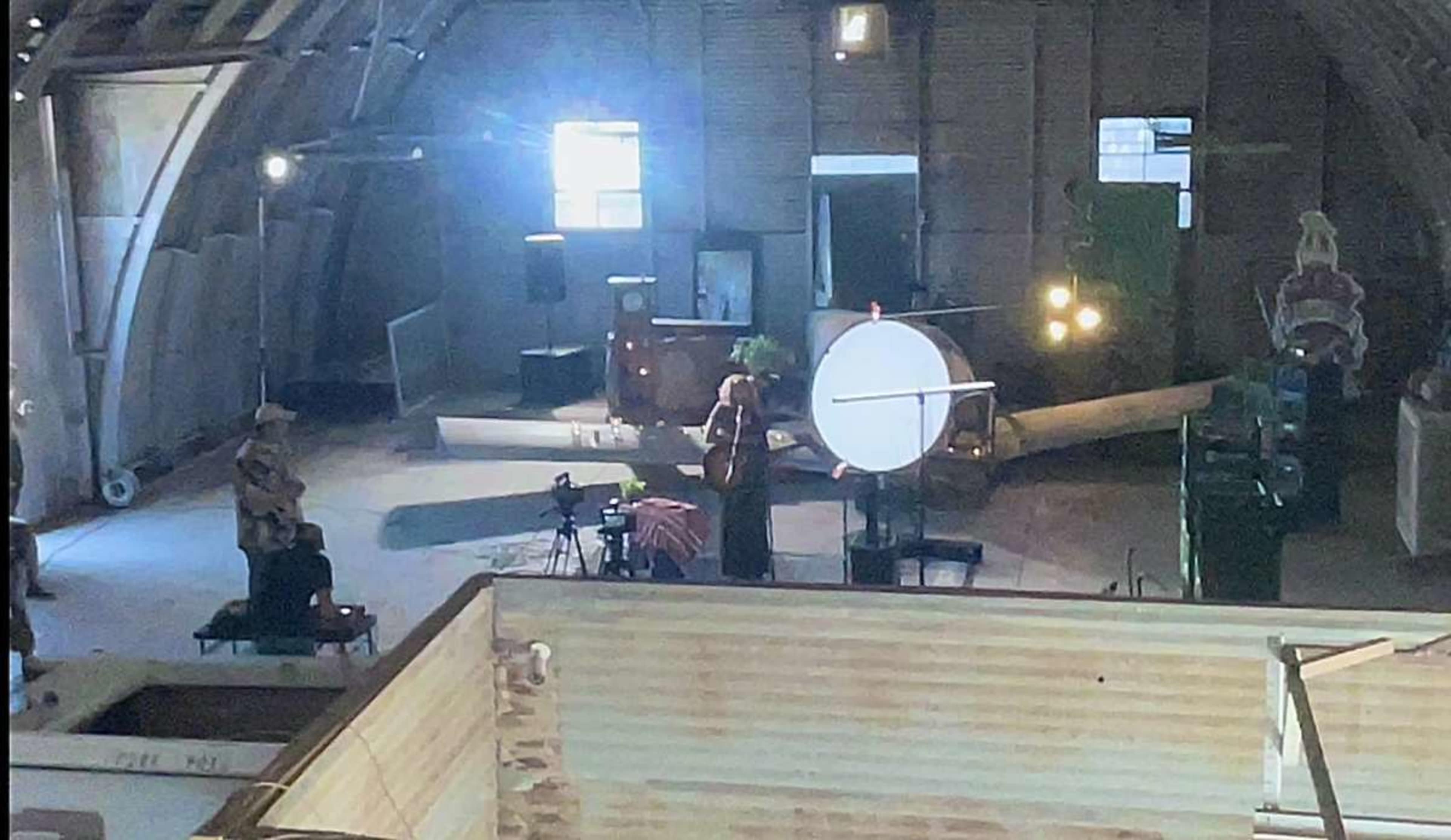 A person stands in an open space with a large circular object while another person sits nearby in a dimly lit, industrial setting.