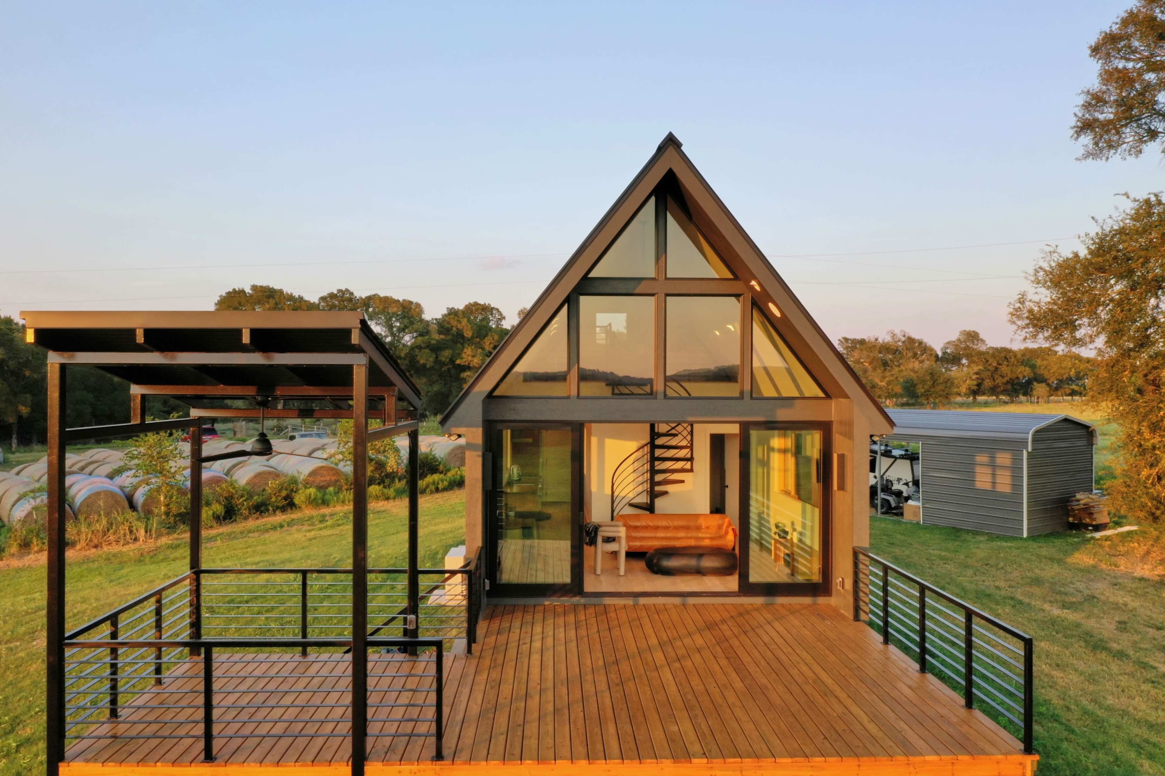 The image depicts a modern A-frame house with large windows, situated on a grassy landscape that includes agricultural storage in the background.