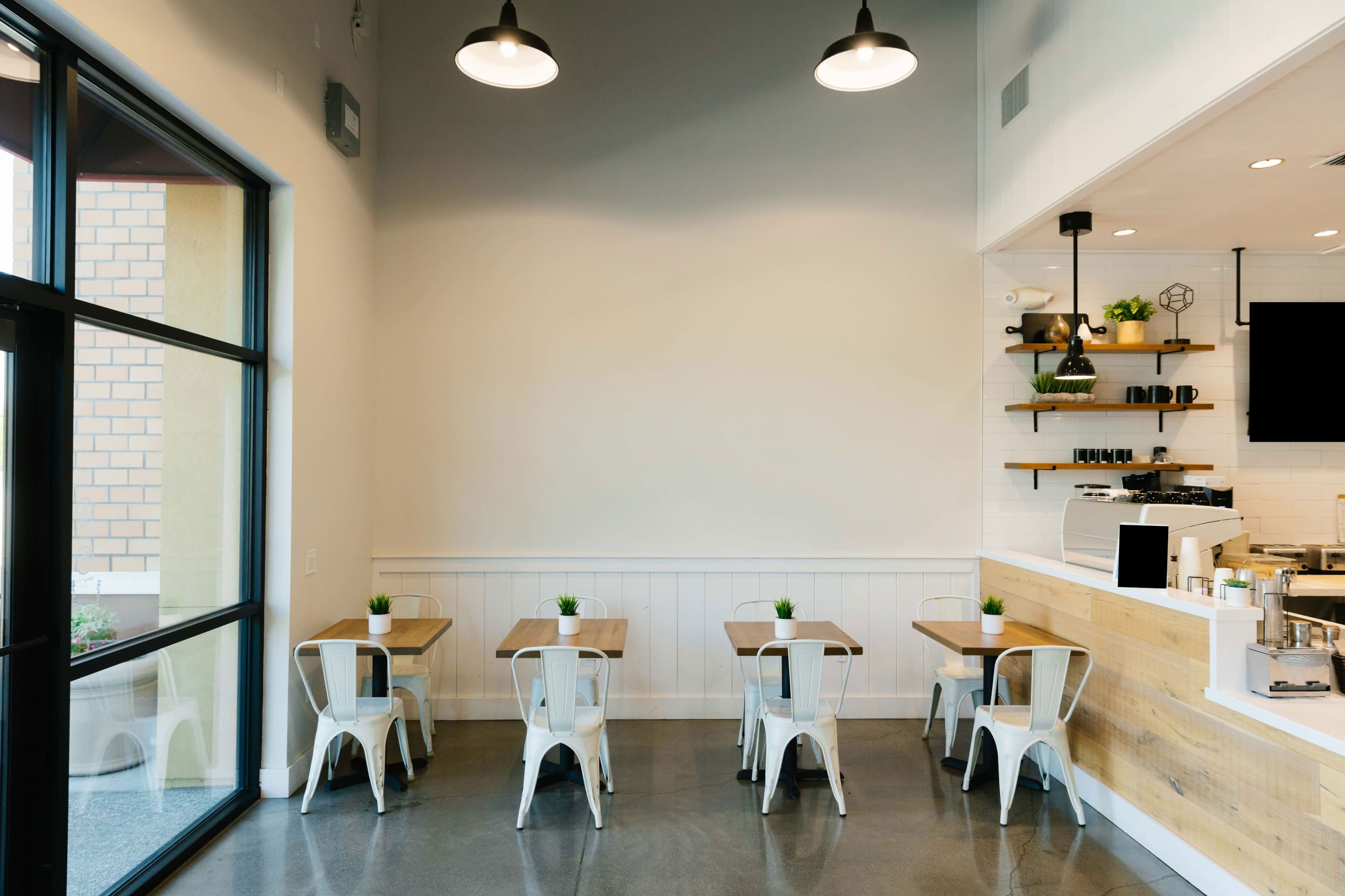 A minimalistic cafe interior with four wooden tables, metal chairs, and a counter area featuring shelves and a coffee machine.
