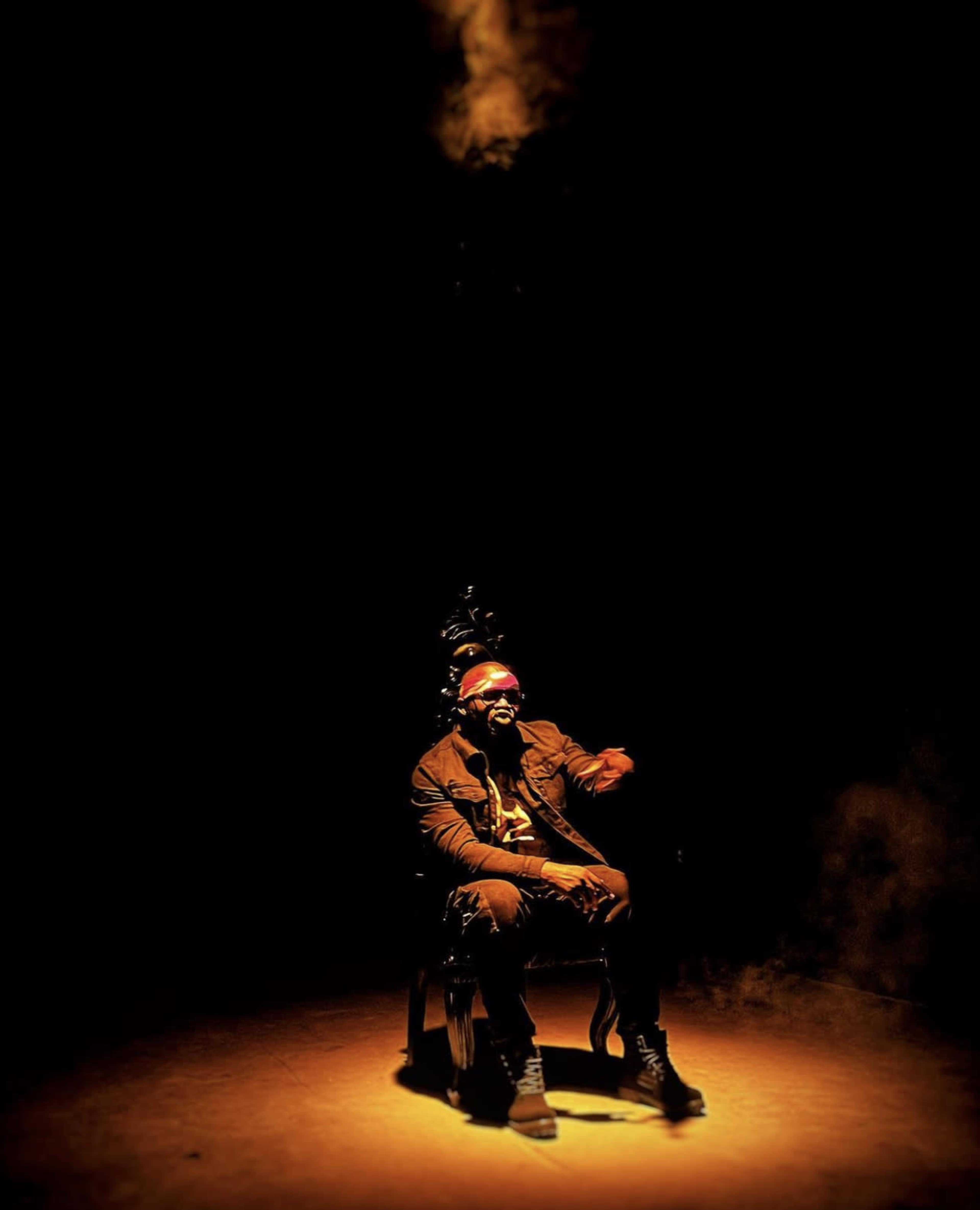 A person sits in a dimly lit room on an ornate chair, with a spotlight illuminating them while smoke swirls overhead.