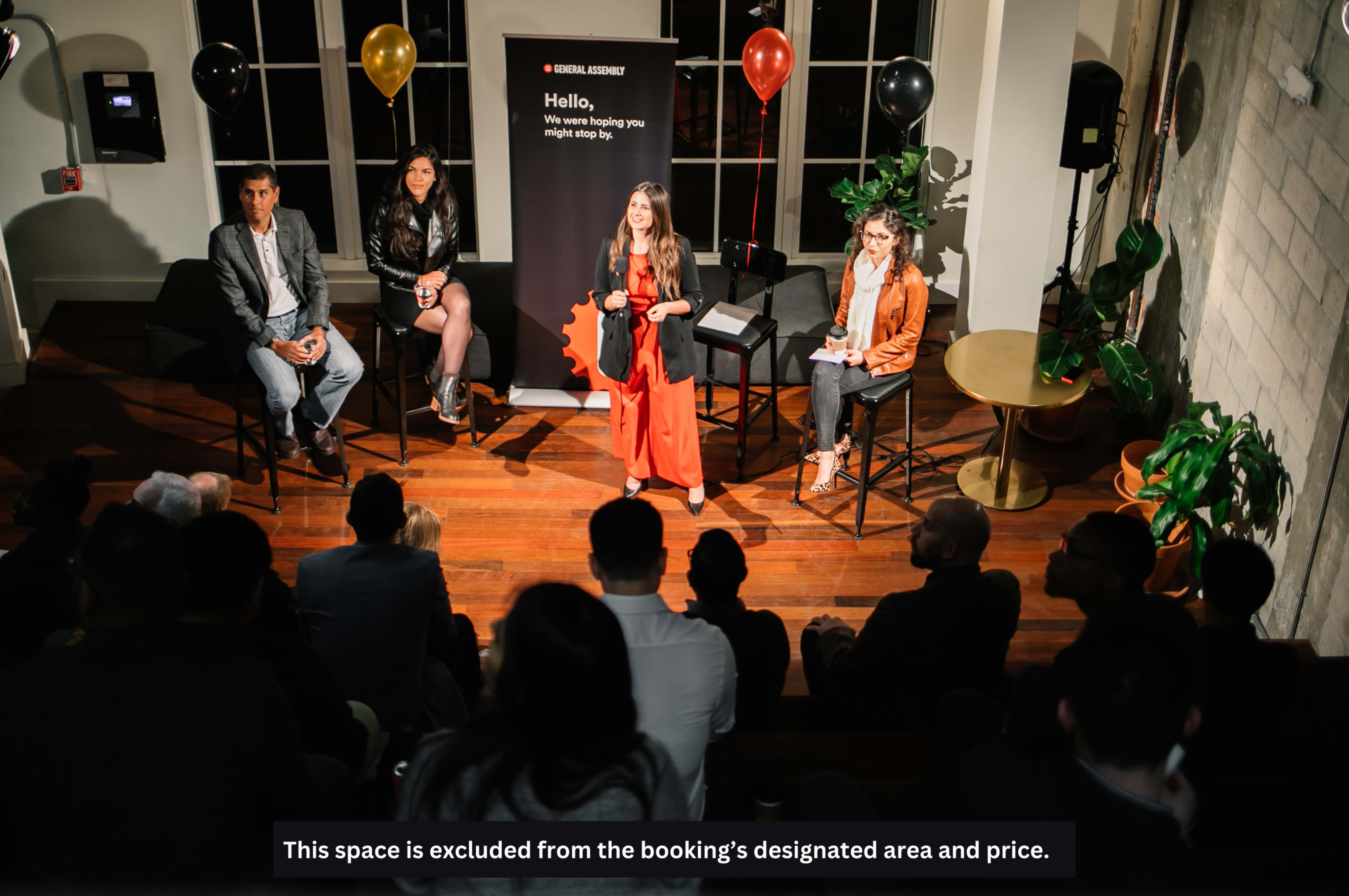 A panel discussion is taking place on stage with three speakers seated on bar stools, while an audience is seated in front of them.