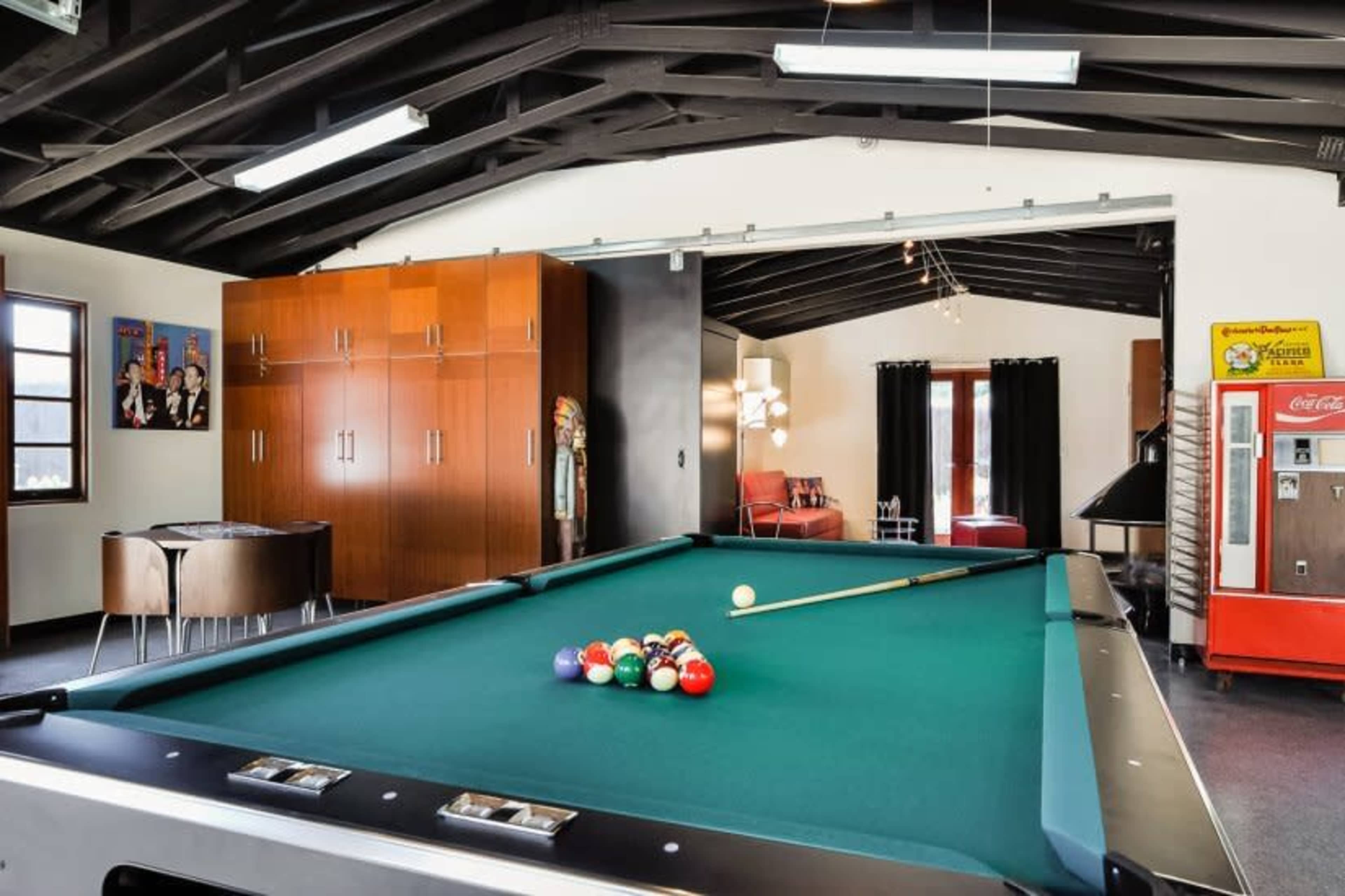 A pool table with a full rack of balls is positioned in a game room featuring high ceilings, a vintage vending machine, and a seating area.