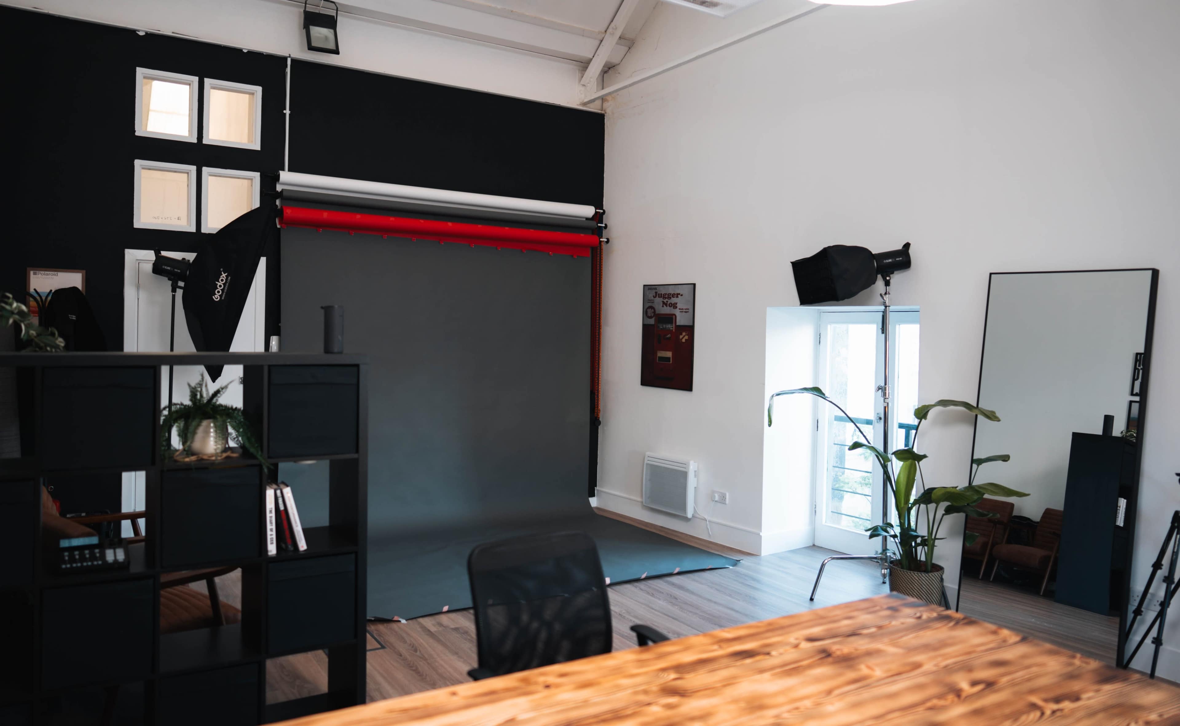 The image shows a modern studio space with a dark backdrop, photography equipment, a large mirror, a desk, and potted plants.