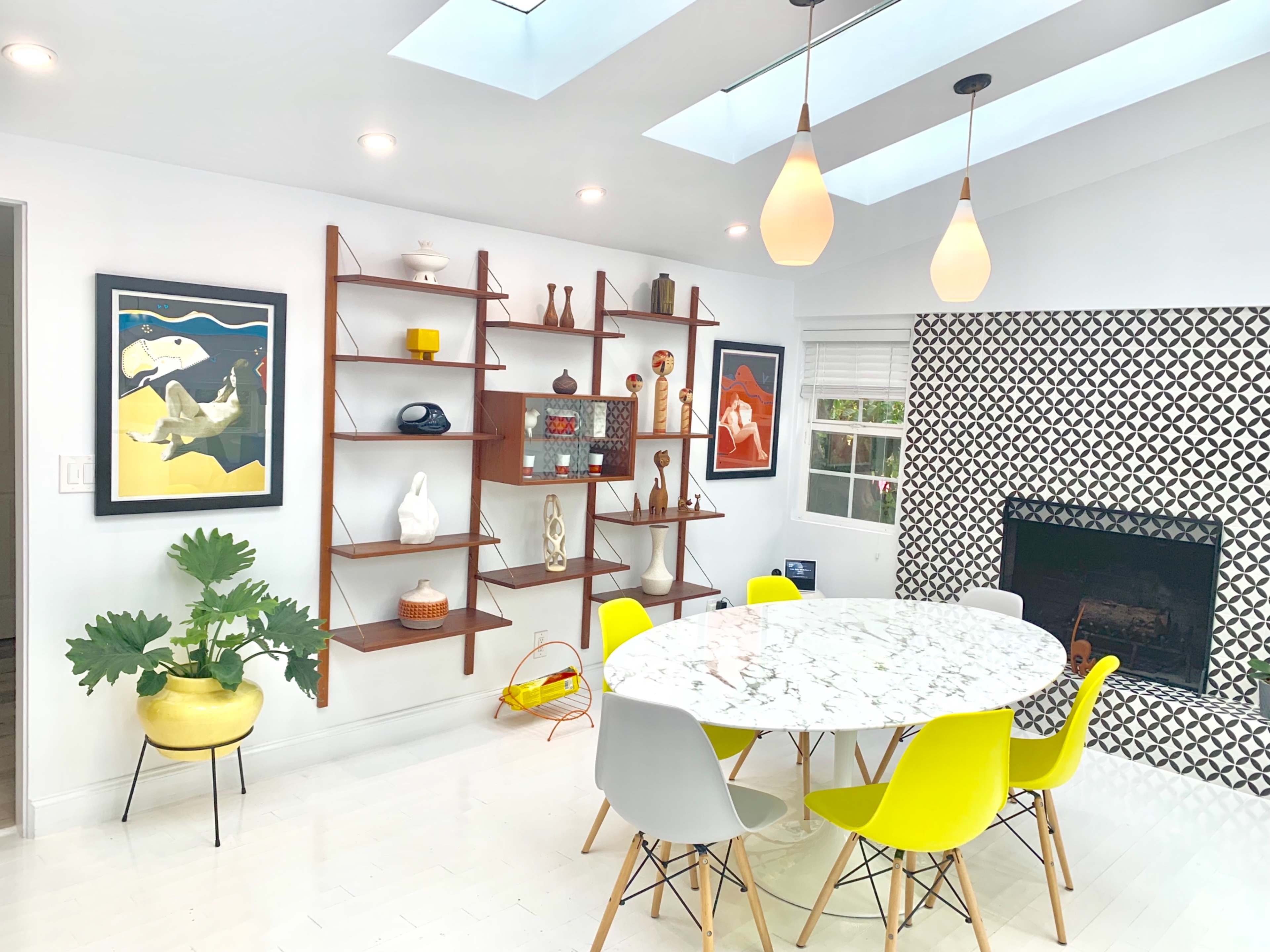 A modern dining area features a round marble table surrounded by colorful chairs, a decorative bookshelf, and a geometric patterned accent wall.