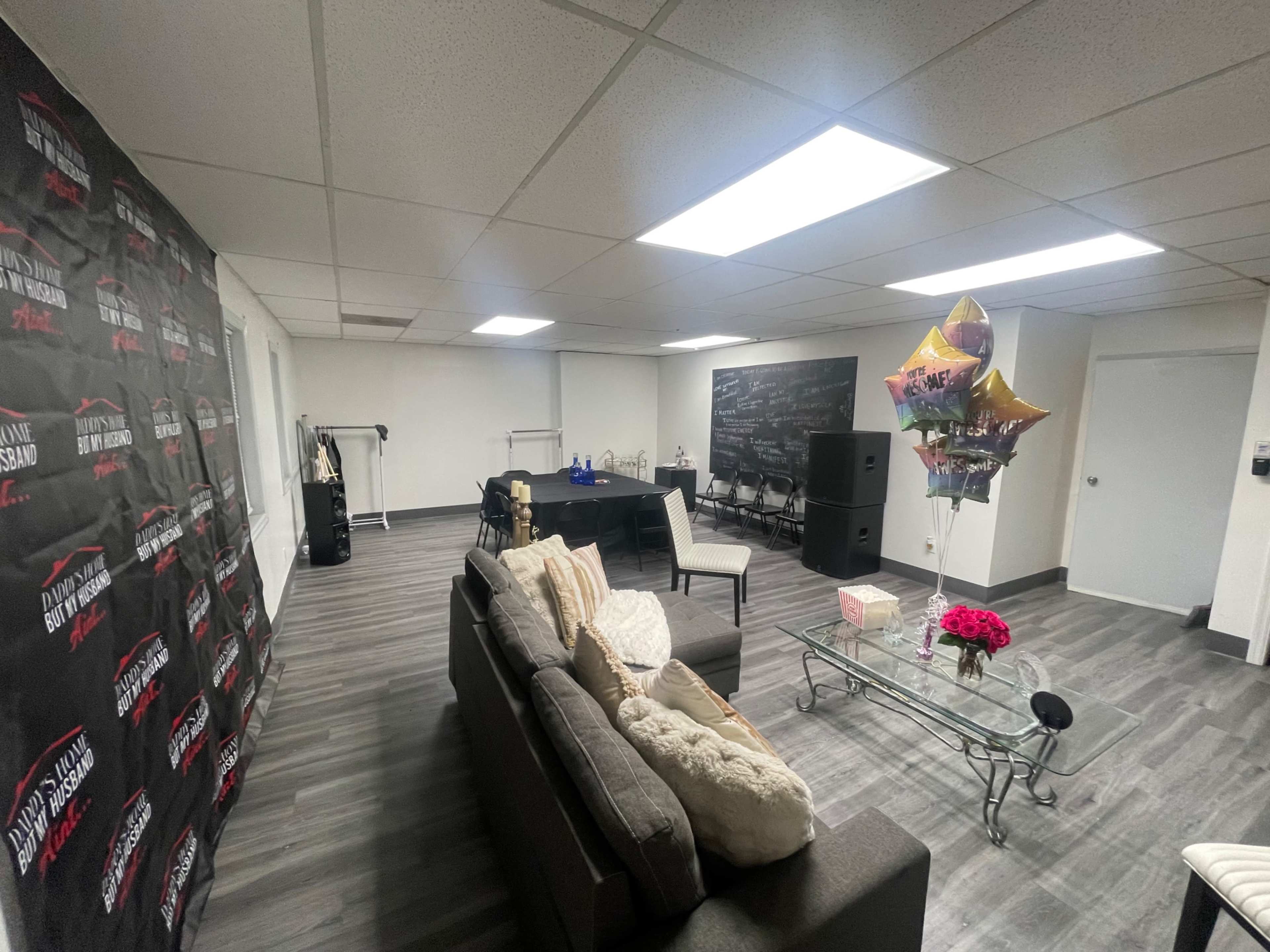 The image depicts a modern, well-lit event space featuring a sofa, a glass coffee table with flowers, and several chairs arranged around a central table.