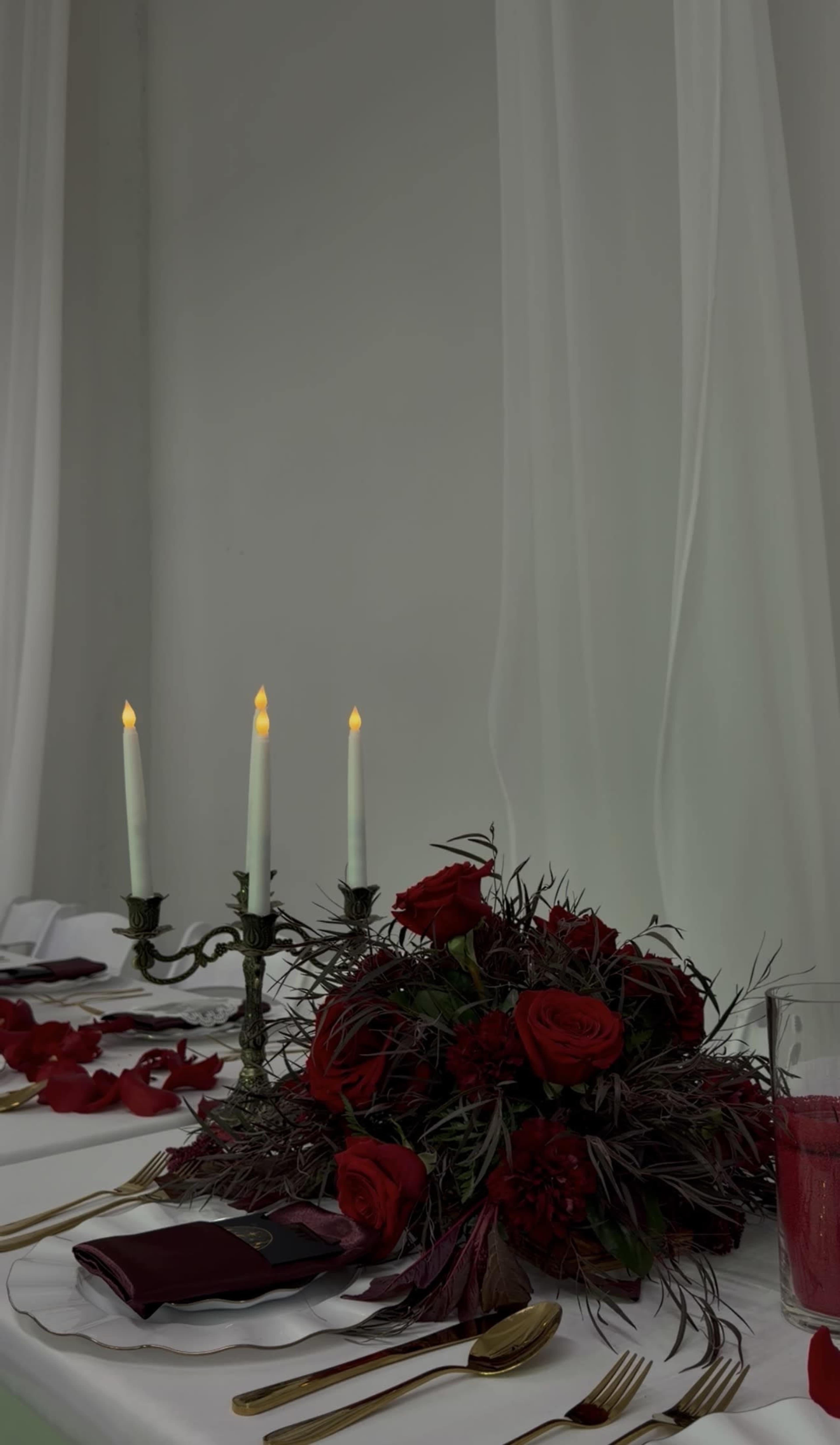 A table is set with a floral centerpiece featuring red roses and greenery, complemented by three lit candles and scattered rose petals.