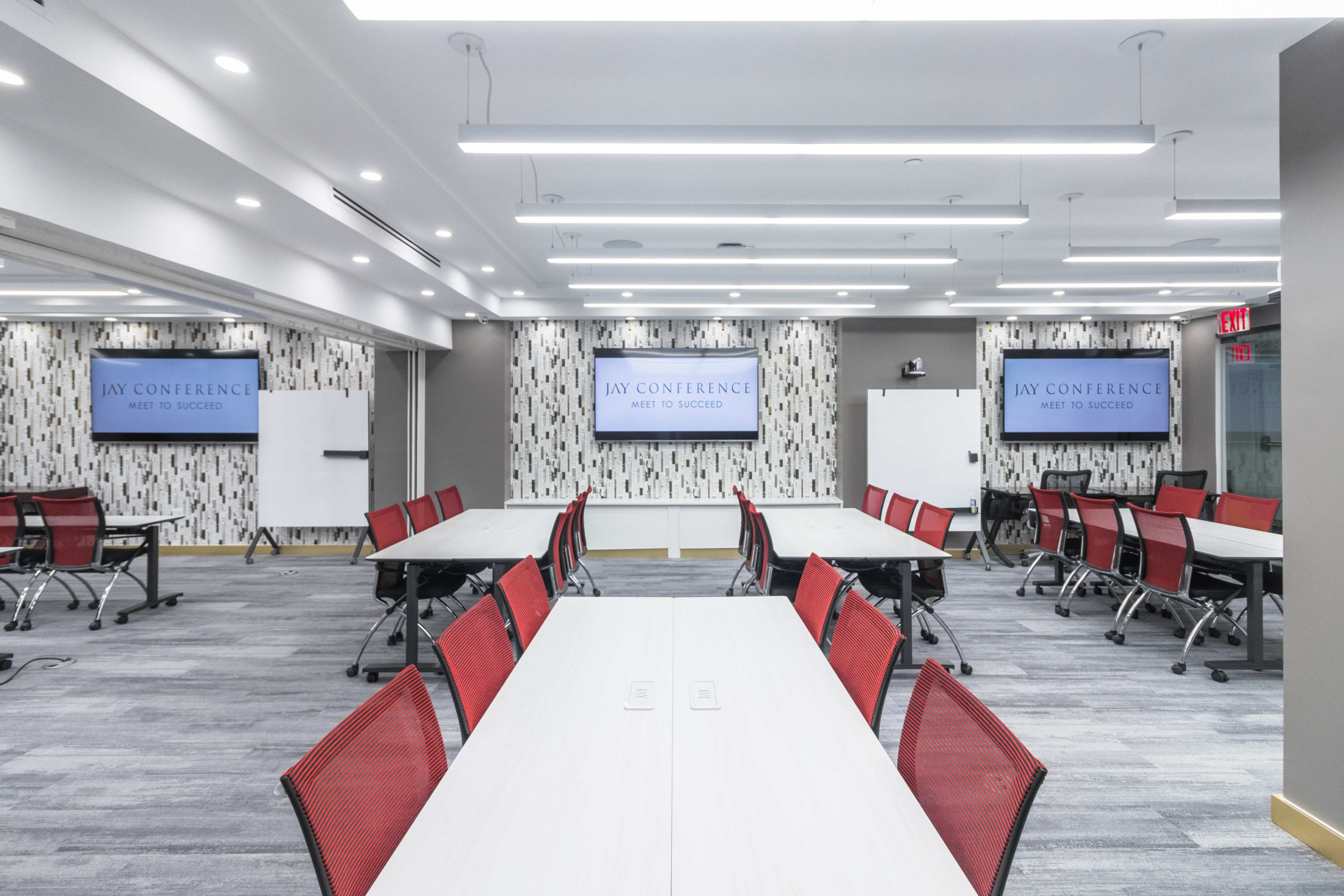 The image shows a modern conference room with multiple tables arranged in a U-shape, featuring red chairs and two large screens displaying "JAY CONFERENCE MEET TO SUCCEED" on the walls.