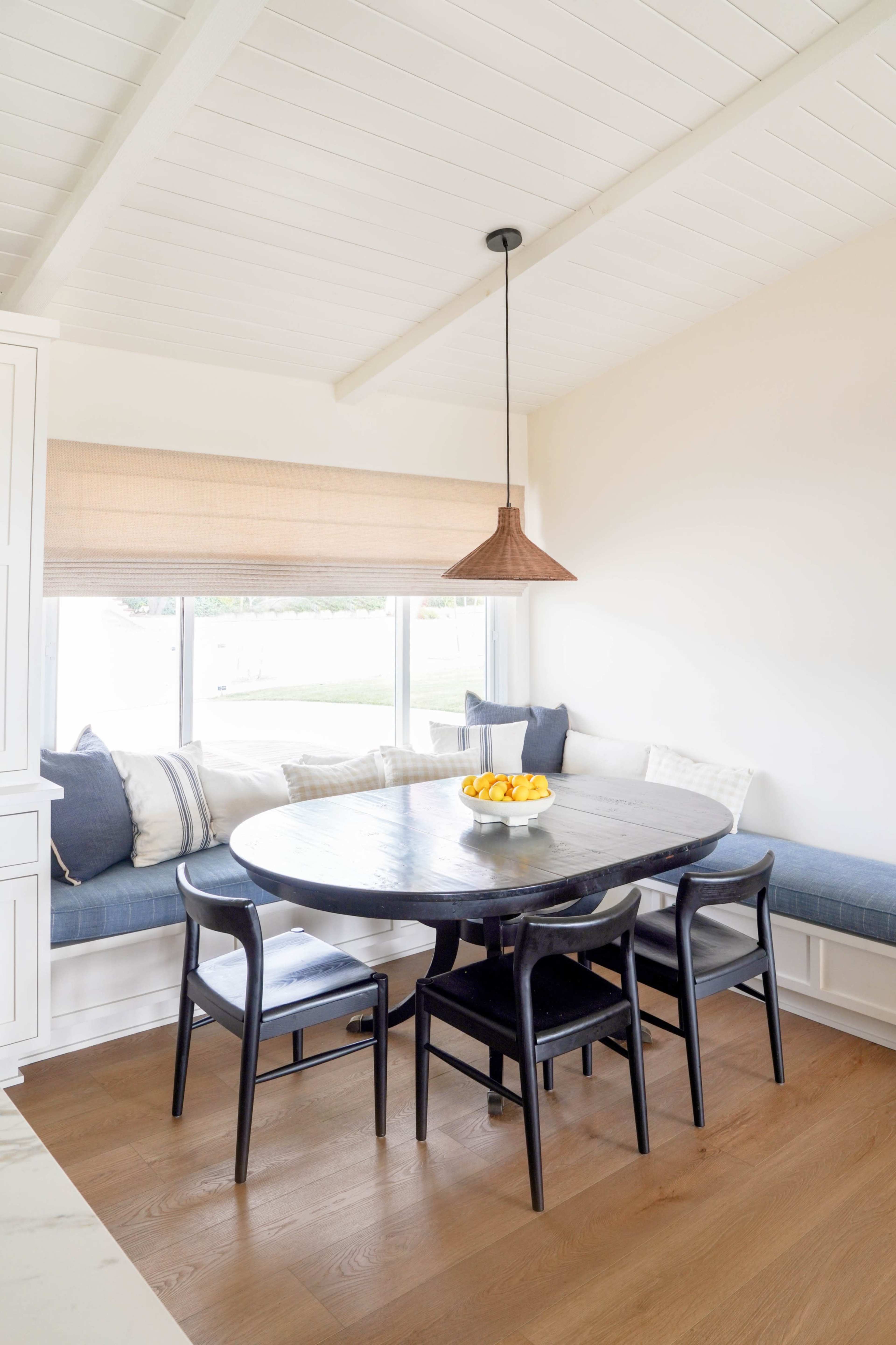 A round dining table with black chairs is positioned near a window, accompanied by a built-in bench and decorative pillows.