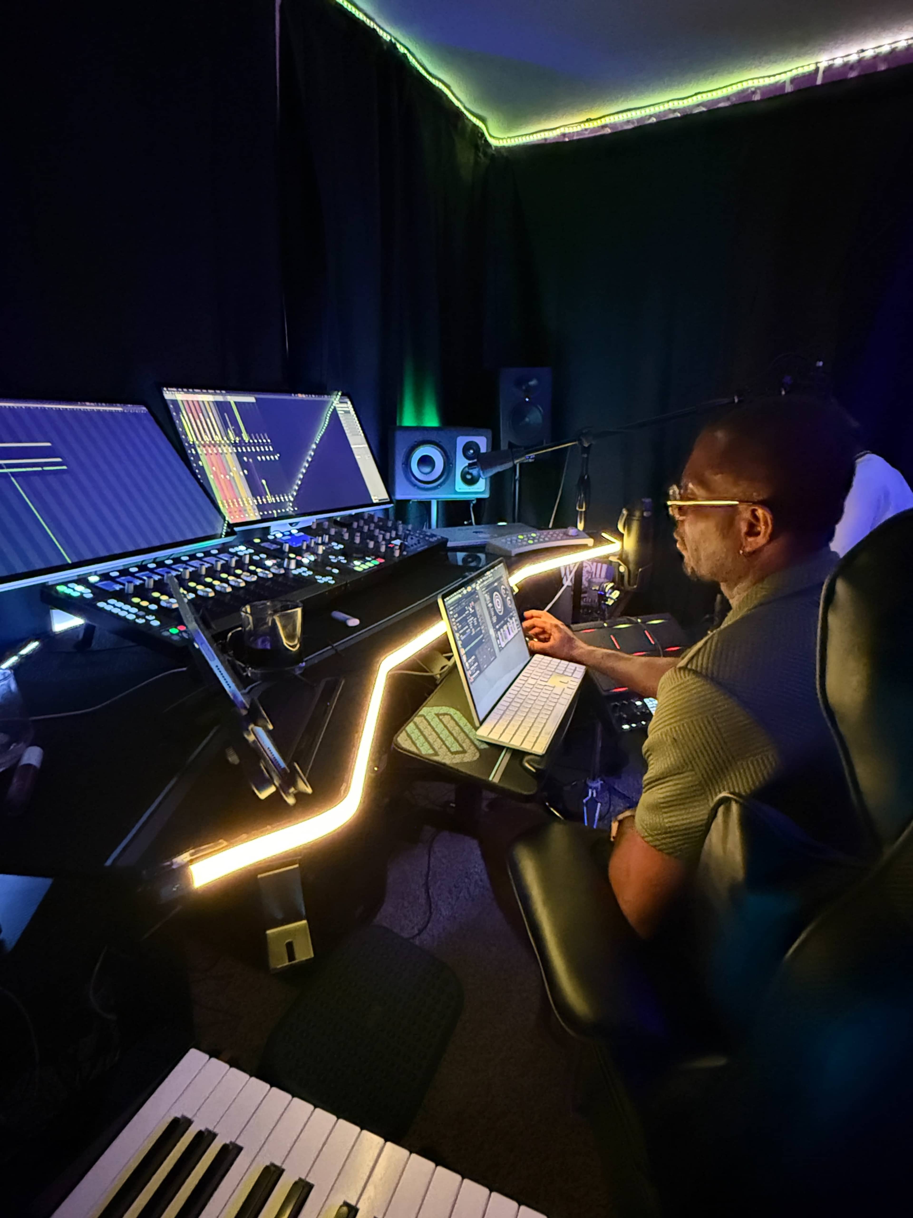 A man sits at a modern music production workstation with multiple screens, a digital audio mixer, and musical equipment in a dimly lit studio.