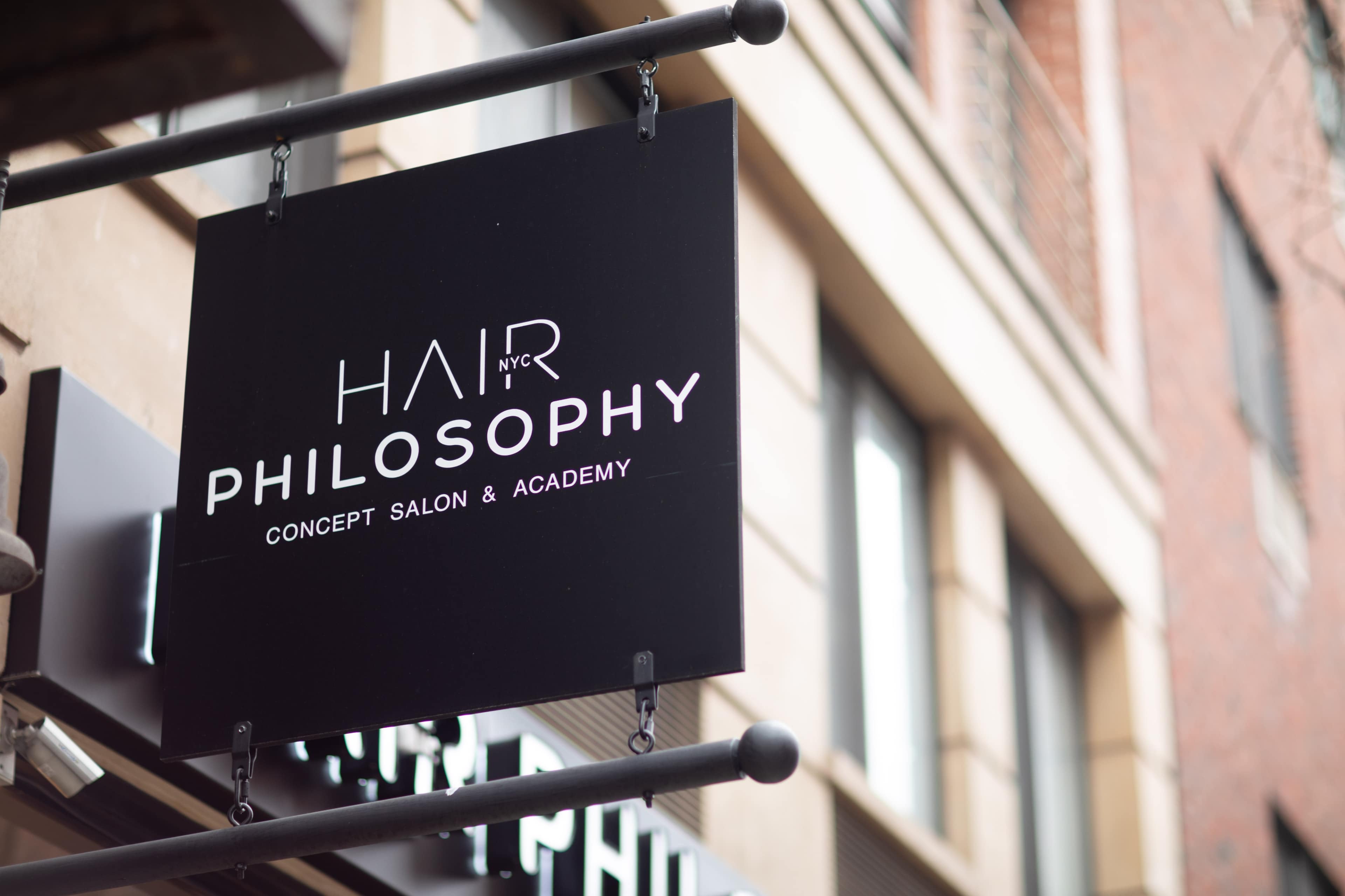 A black sign hanging outside a building, displaying the text "HAIR PHILOSOPHY CONCEPT SALON & ACADEMY."