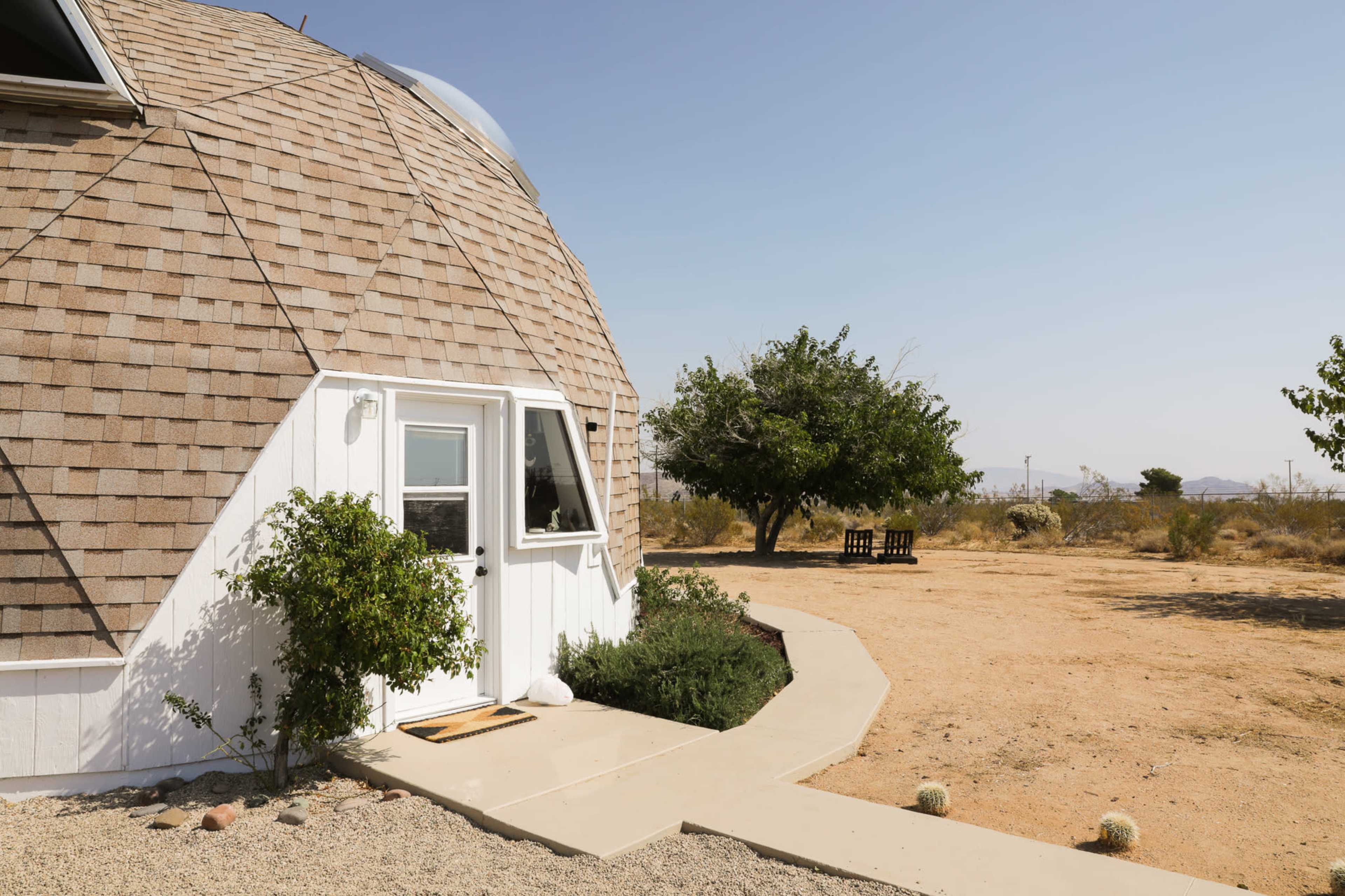 A geodesic dome house with a shingled roof sits on a sandy lot surrounded by sparse greenery and desert landscape.