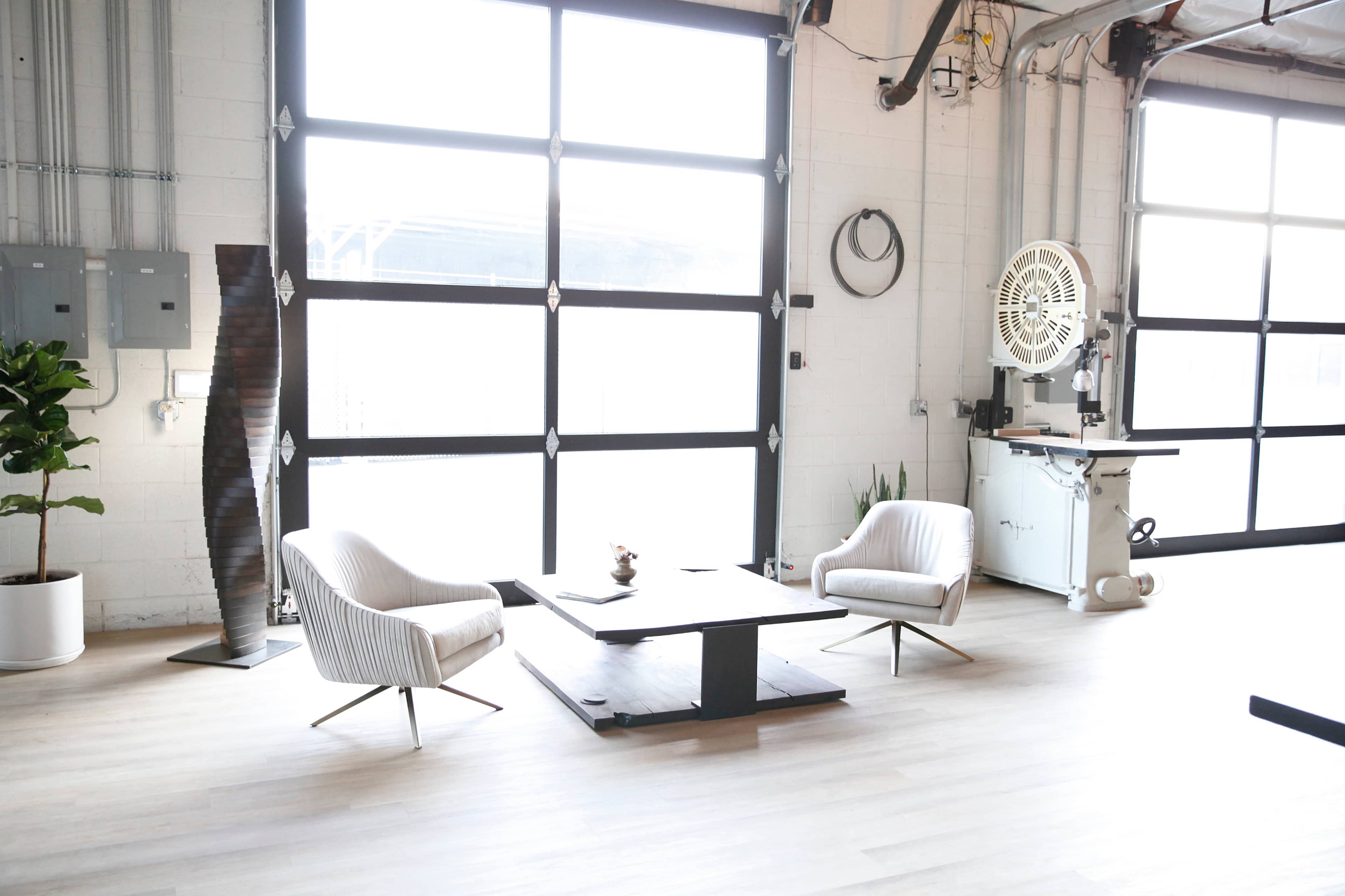 A modern interior space with two striped armchairs facing a low wooden coffee table, a plant in a white pot, and large windows allowing natural light.