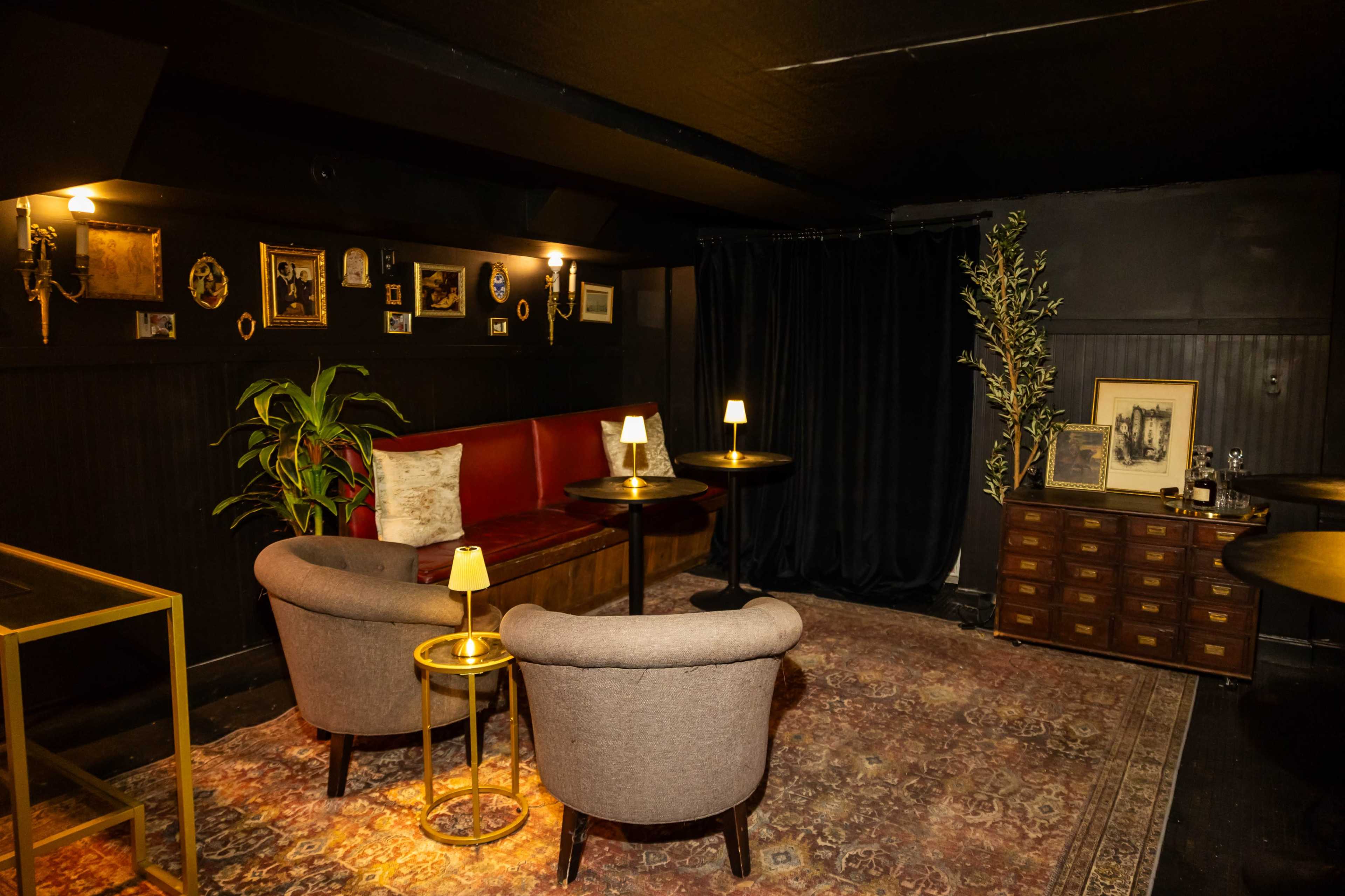 The image shows a dimly lit lounge area featuring a red upholstered bench, two armchairs, small tables with lamps, and framed photographs on the walls.