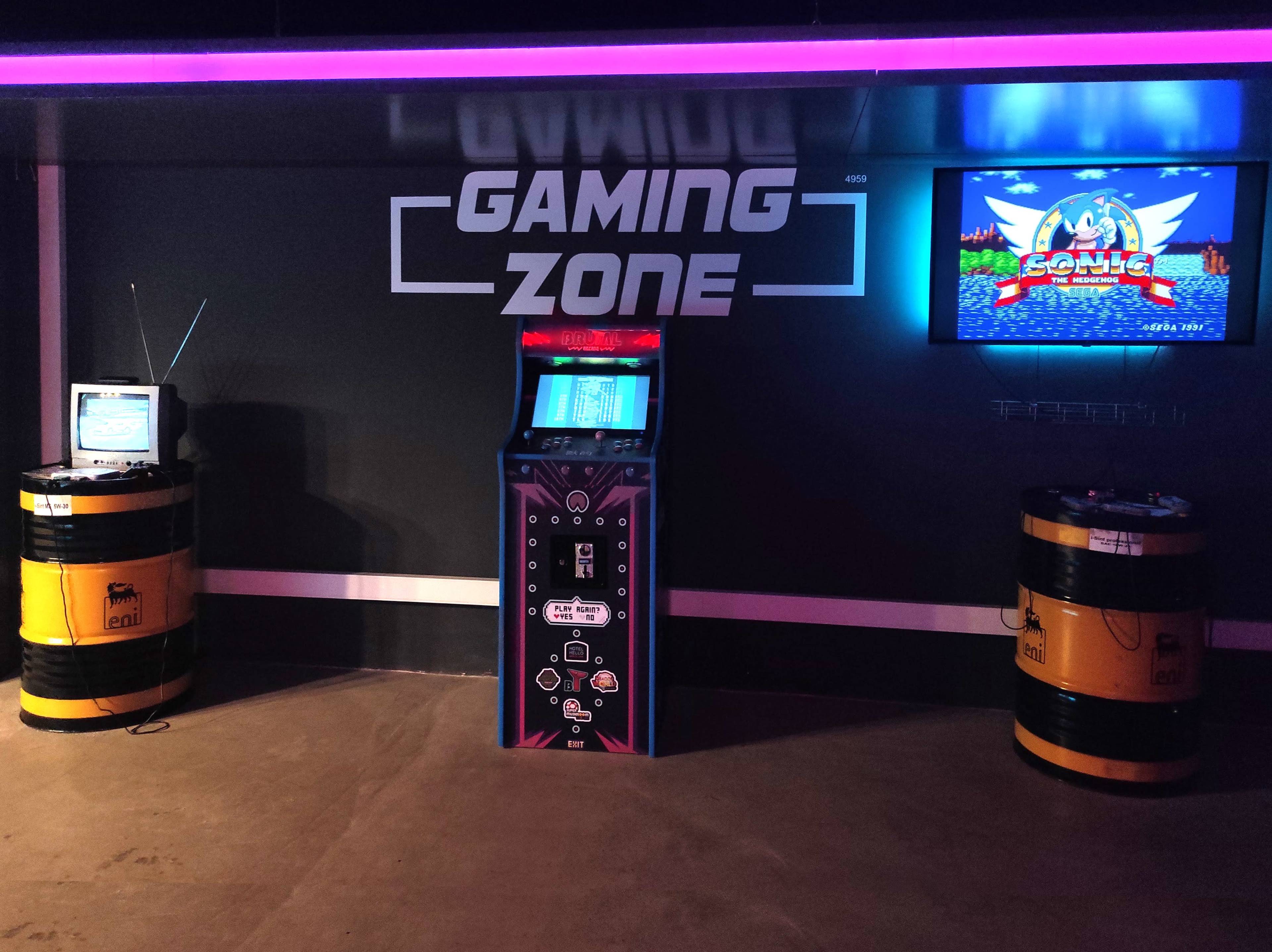 A gaming zone featuring an arcade machine in the center, flanked by a vintage television on the left and a modern screen displaying a Sonic game on the right, with yellow barrels as decor.