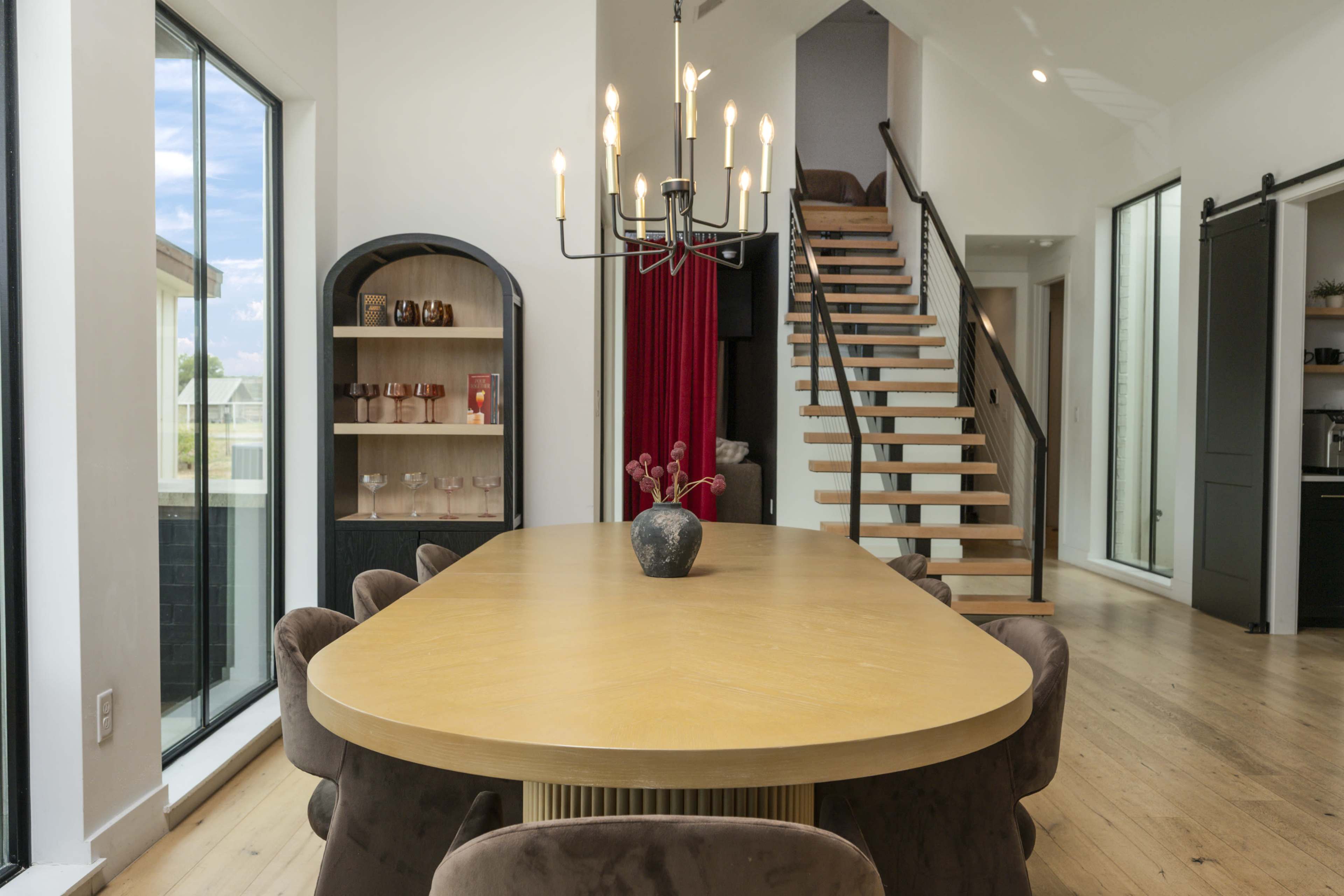 A modern dining space features a long wooden table surrounded by plush chairs, a chandelier overhead, and a staircase leading to an upper level.