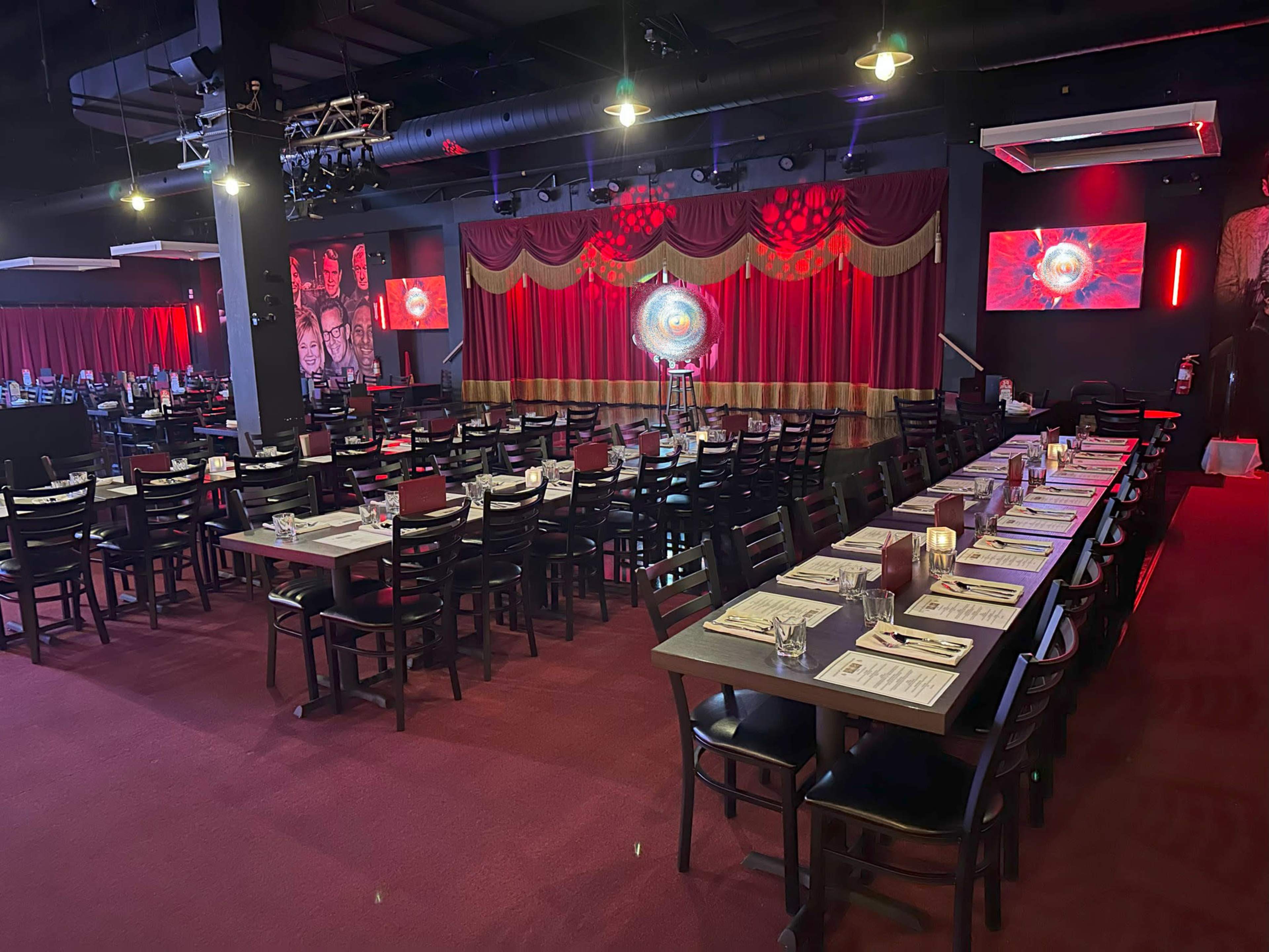 An elegantly arranged restaurant interior featuring rows of tables set for dining, red drapes on the stage, and colorful lighting.