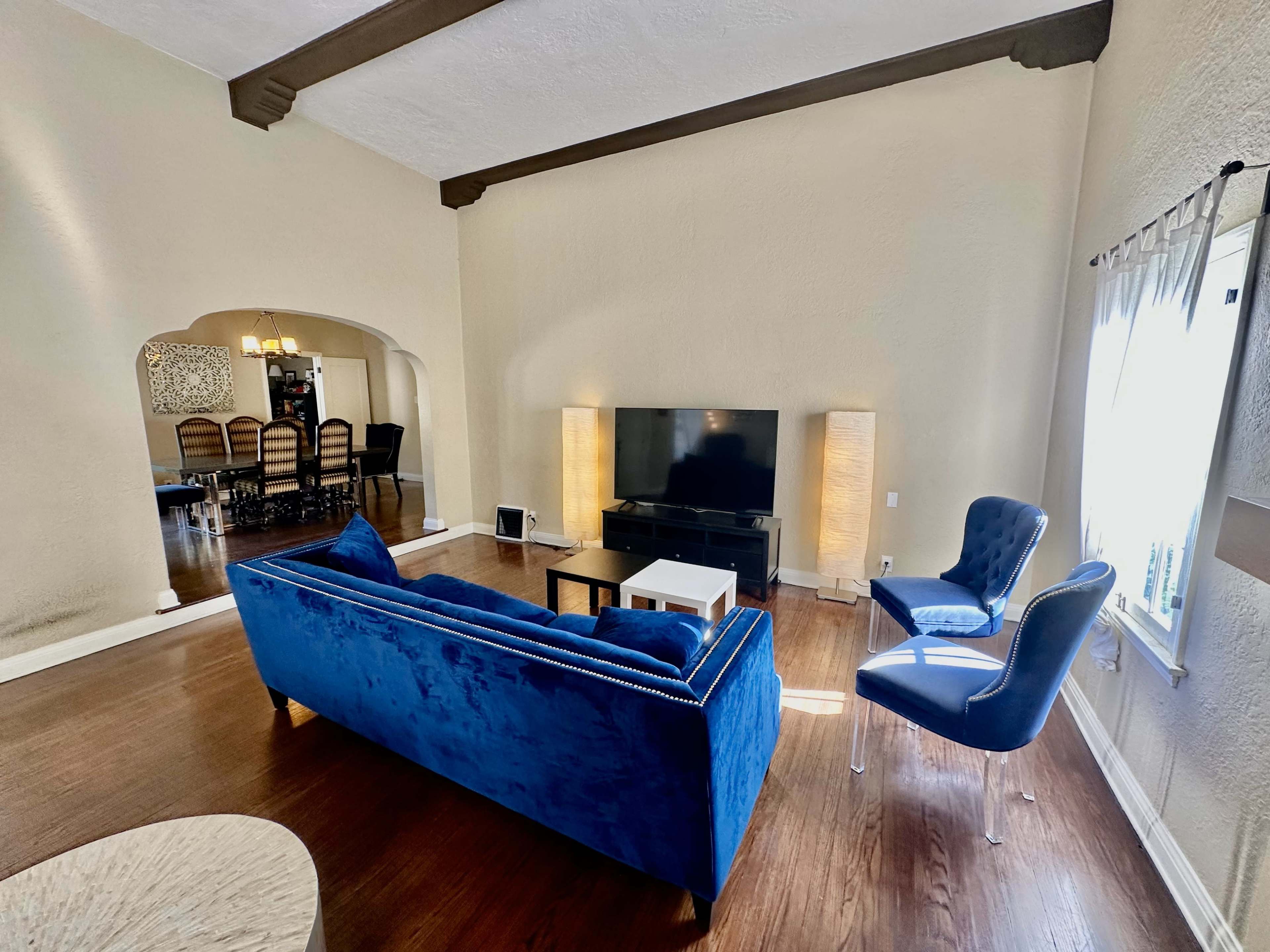 A modern living room features a blue velvet sofa, a black television stand, and two floor lamps, with a dining area visible in the background.