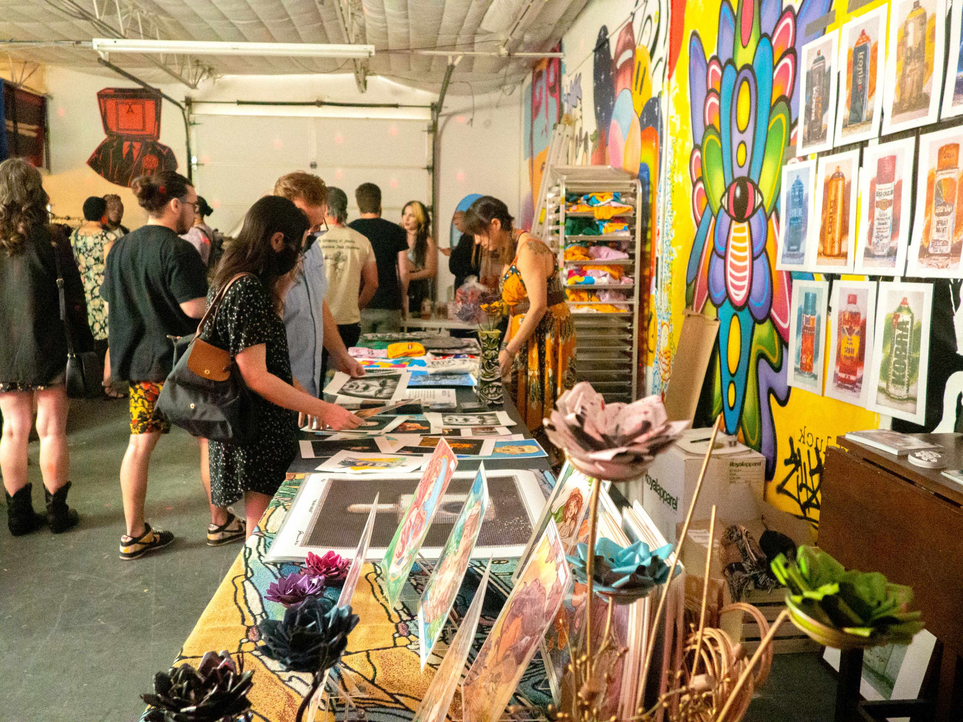 A group of people gathers in an art space, browsing various artworks and prints displayed on tables and walls adorned with colorful murals.