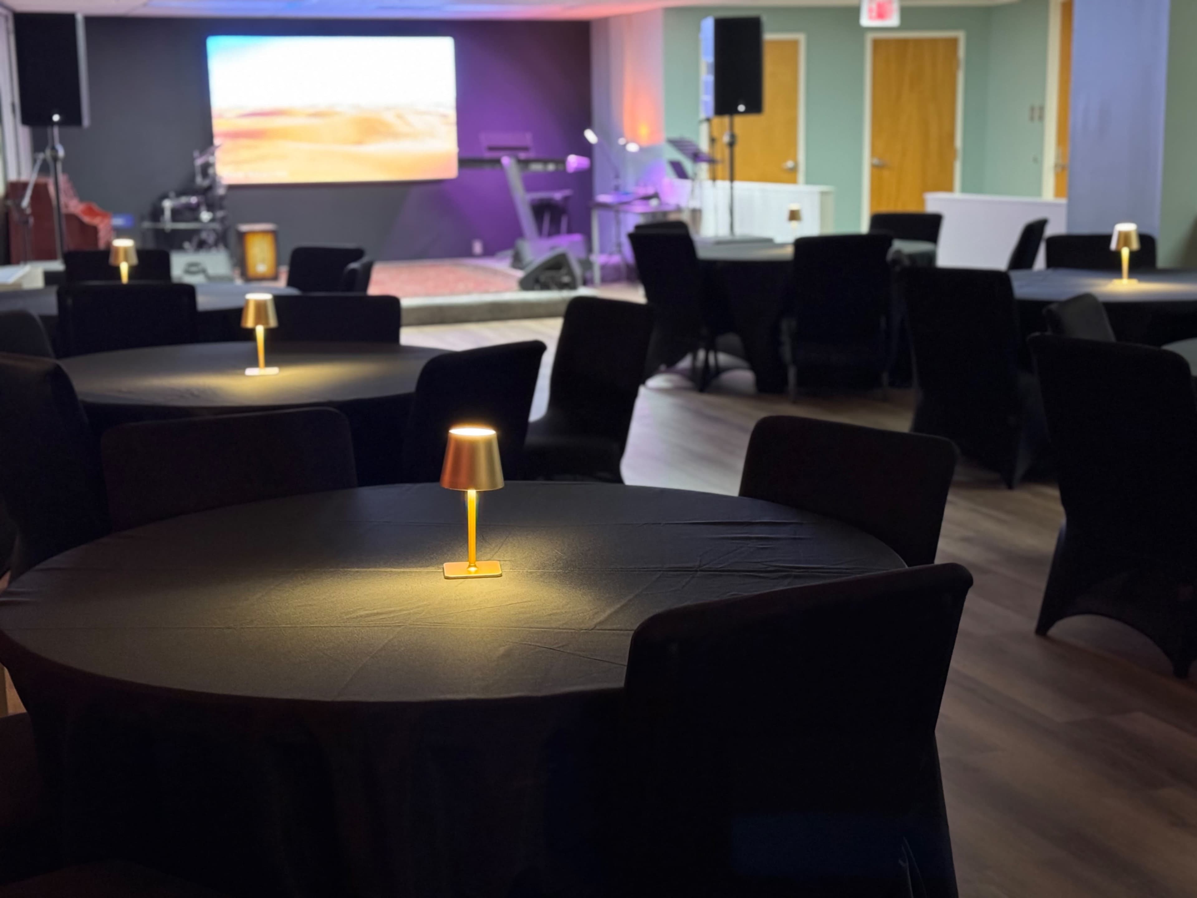 The image shows a modern event space with several round tables covered in black tablecloths, each featuring a small lamp, and a presentation screen and stage area in the background.