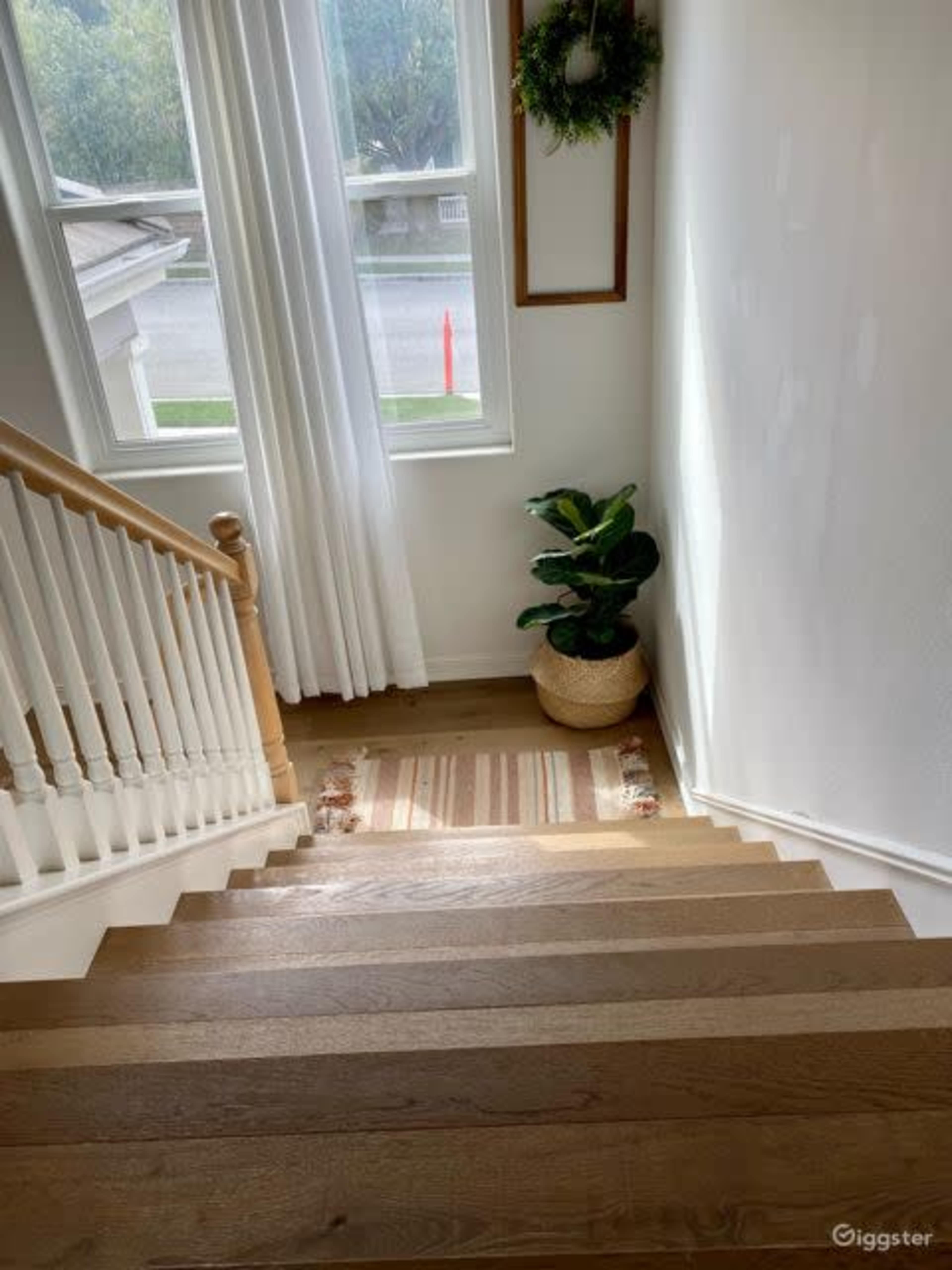 The image shows a staircase leading down to a well-lit entryway with a potted plant and a rug on the floor.