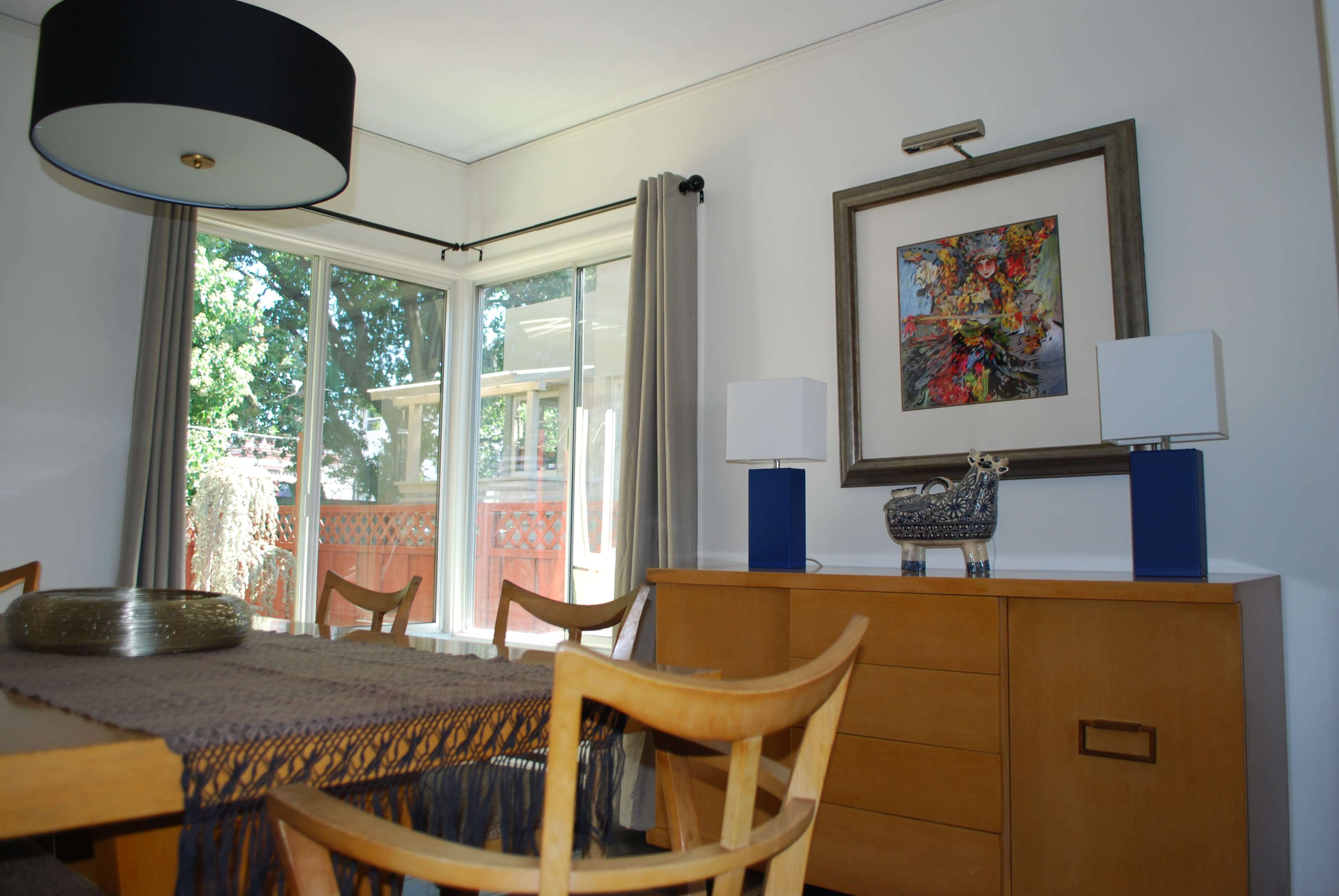 A dining area features a wooden table, four chairs, a light fixture, and a sideboard with decorative items, alongside large windows letting in natural light.