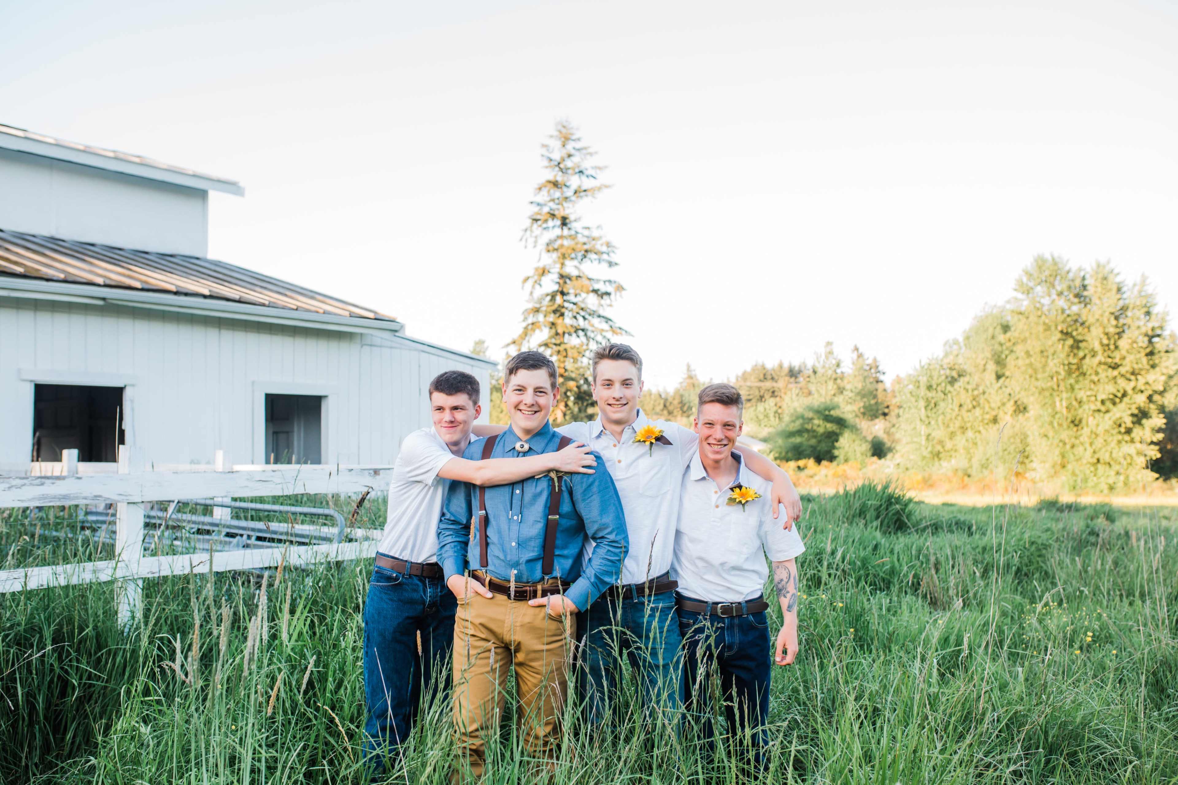 Four young men stand together in a field, smiling, with a white barn and trees in the background.