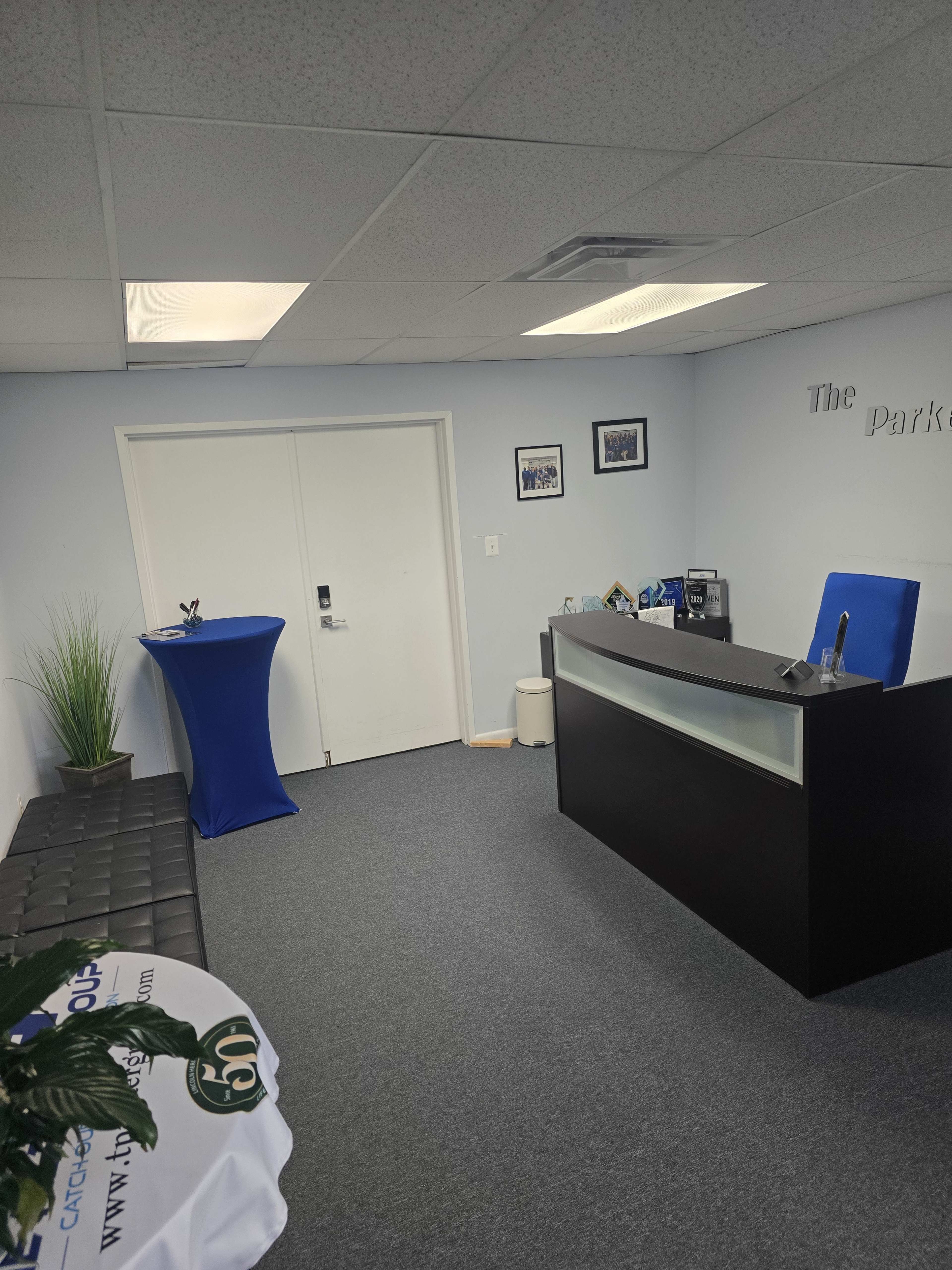The image shows a modern office reception area with a black front desk, a blue tablecloth on a table, framed pictures on the wall, and a plant in the corner.