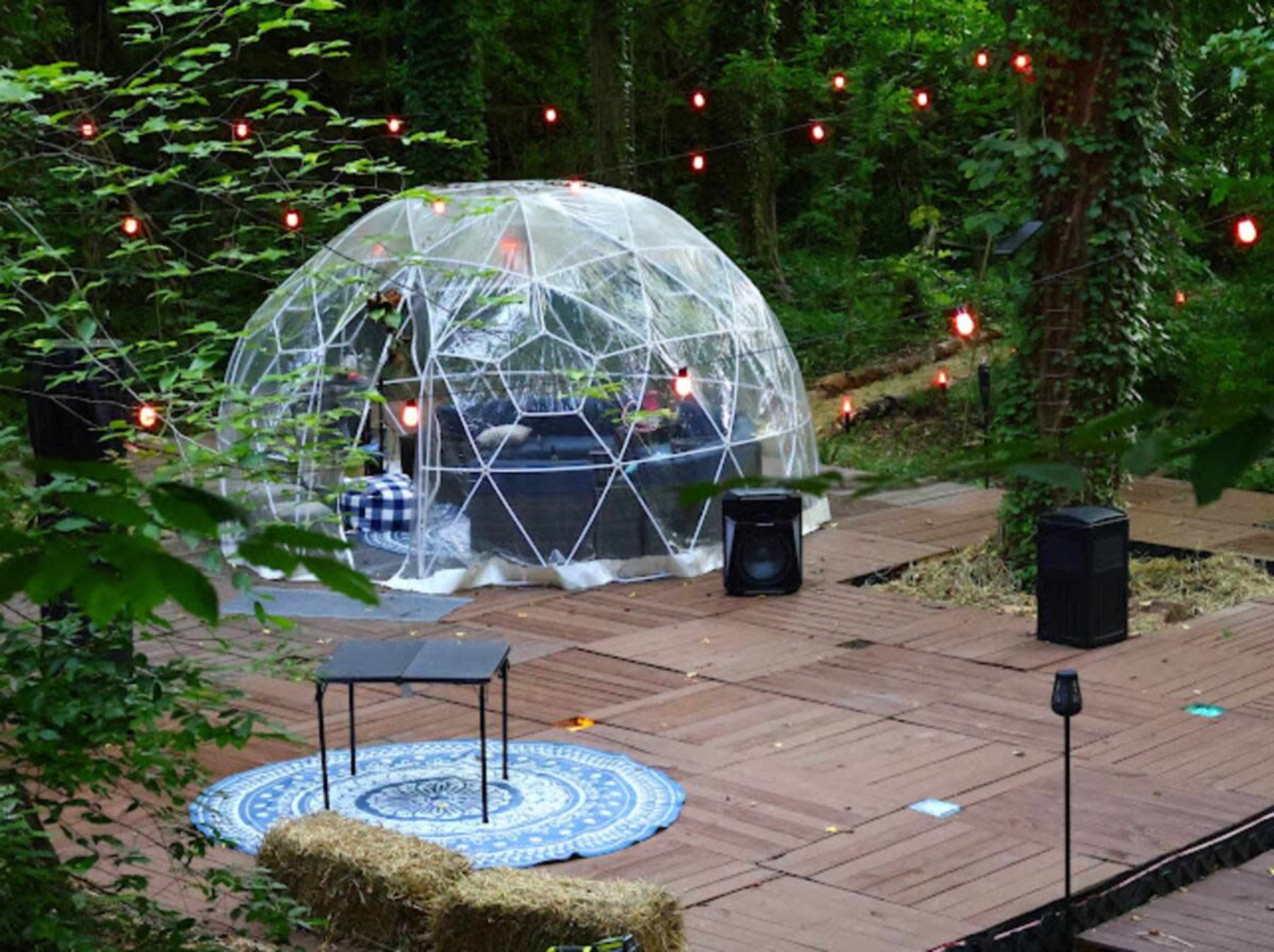 The image shows a transparent geodesic dome on a wooden deck in a lush green forest, surrounded by string lights and a patterned rug.