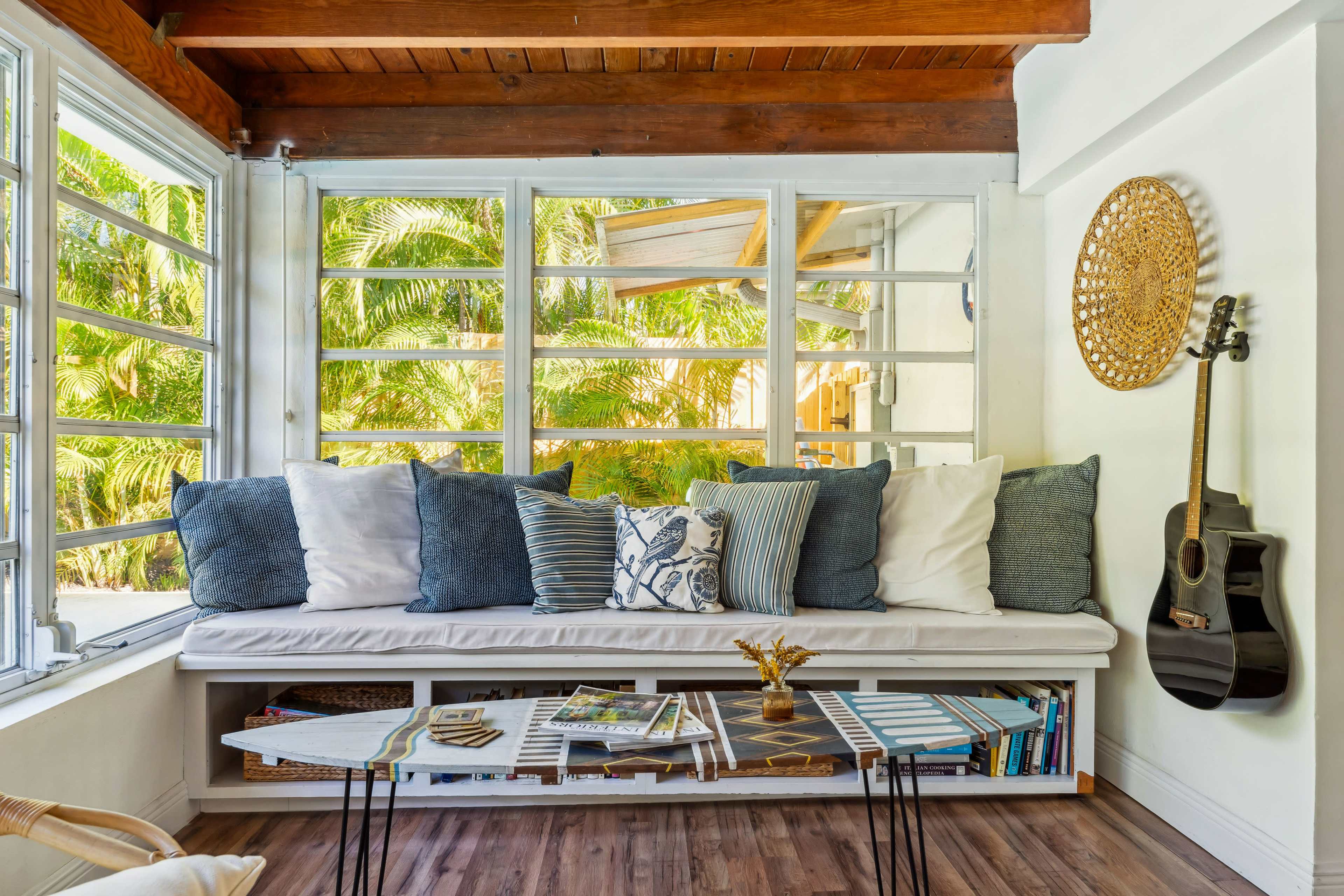 The image shows a cozy seating area with a long cushioned bench, decorative pillows, a coffee table with books, and a guitar leaning against the wall, surrounded by large windows that let in natural light and views of greenery.