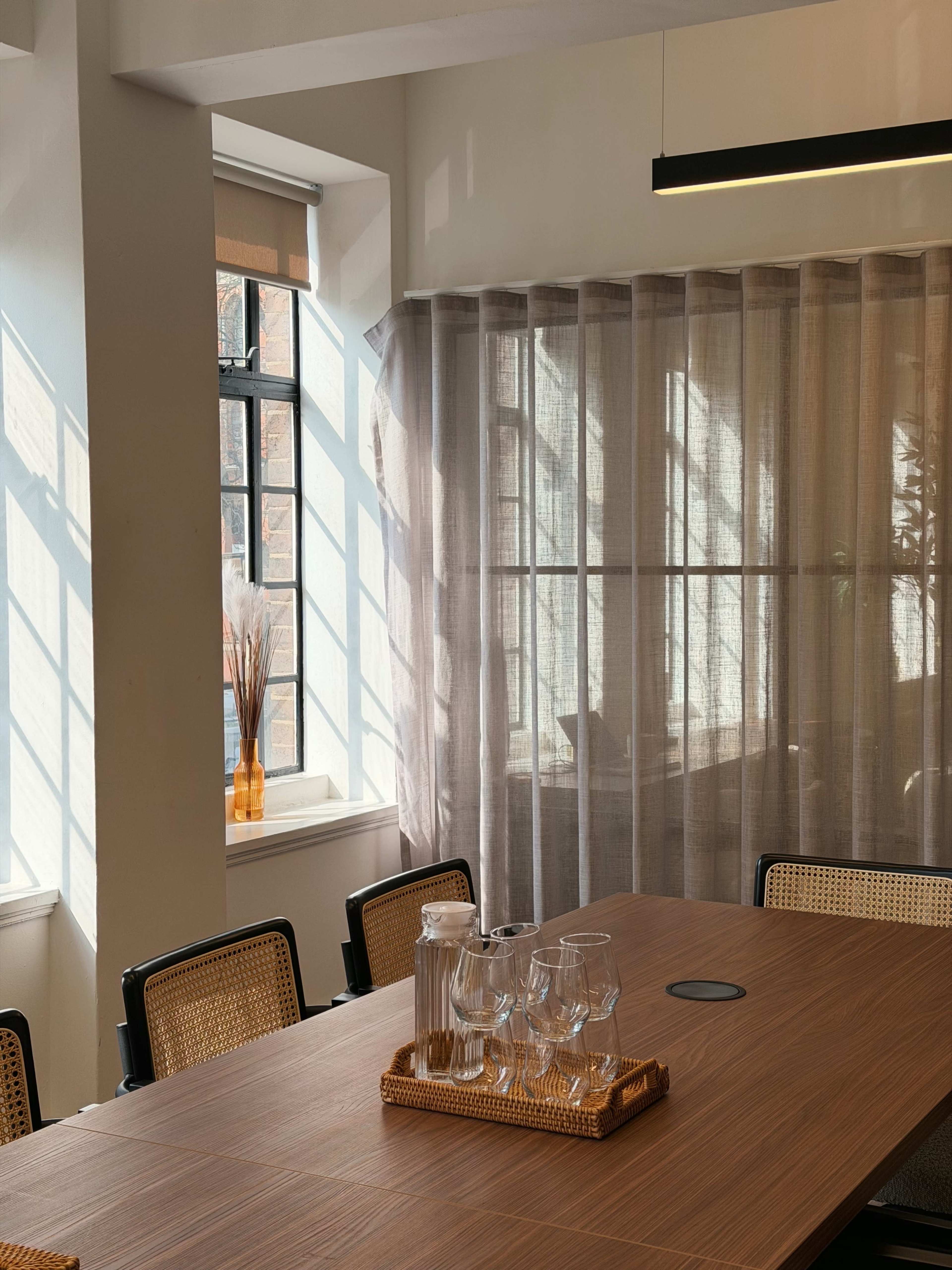 A modern conference room features a wooden table set with glasses and a carafe, illuminated by natural light filtering through large windows.