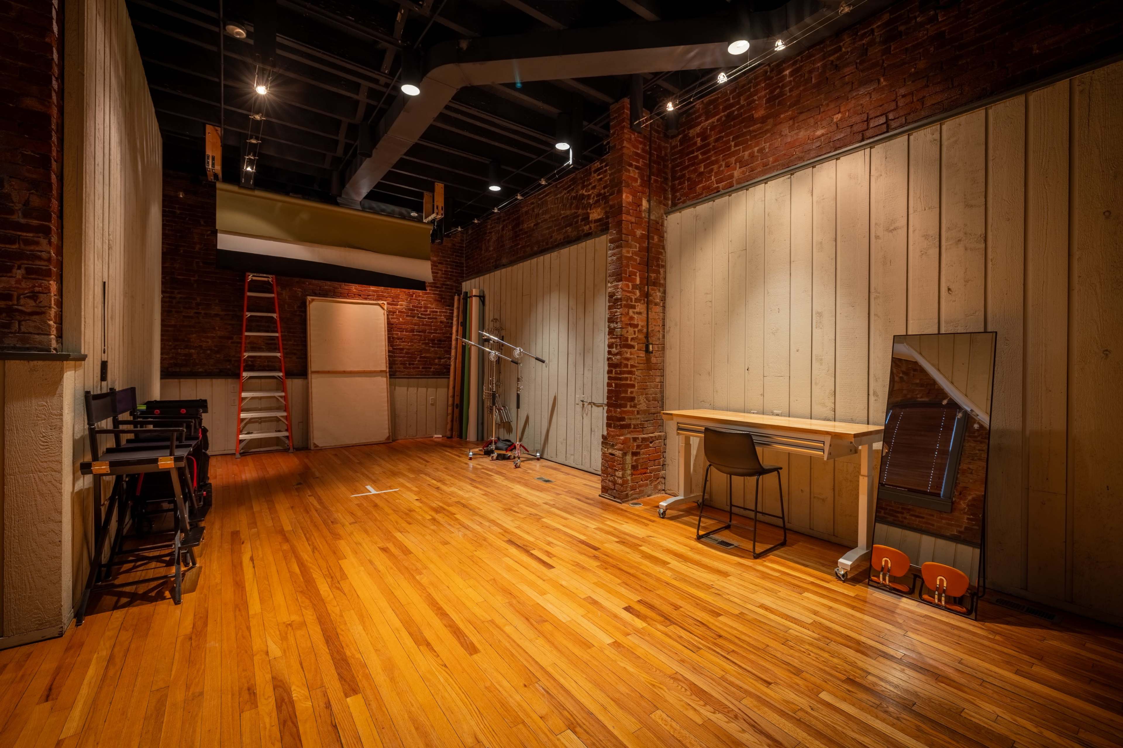 The image shows an empty studio space with exposed brick walls, wooden flooring, and a desk with a chair on one side, along with a ladder and various equipment in the background.