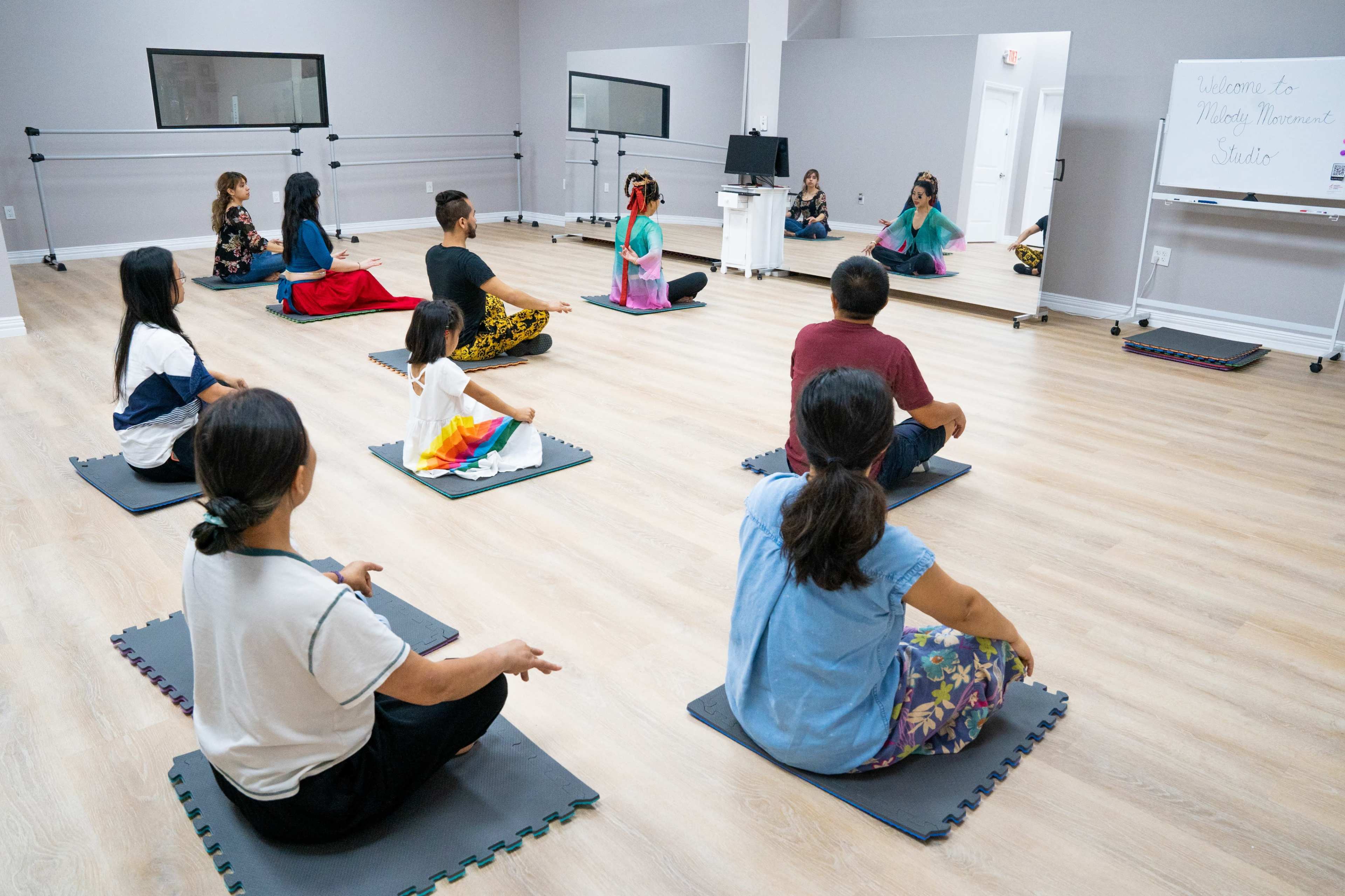 A group of people sits on exercise mats in a dance studio while listening to an instructor.