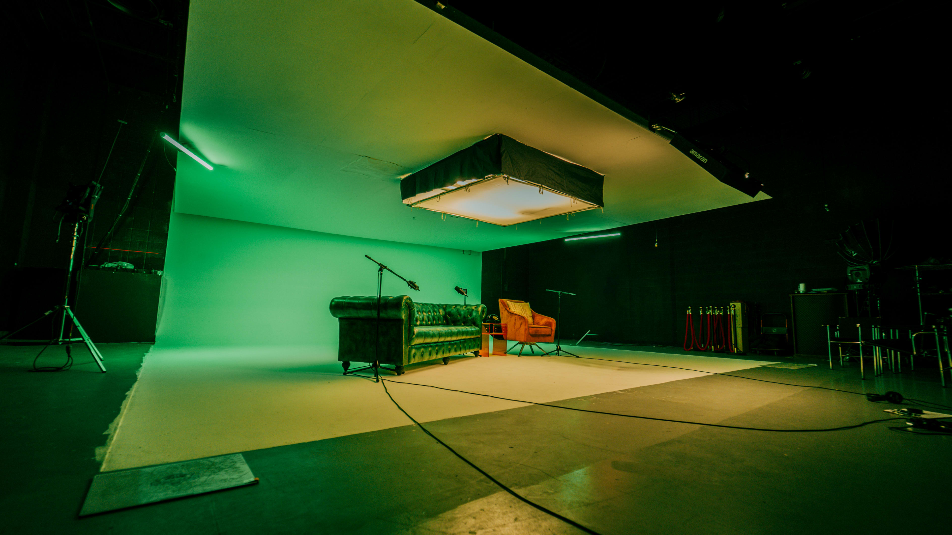 A green screen studio features a green backdrop, a dark green sofa, an orange chair, and overhead lighting.