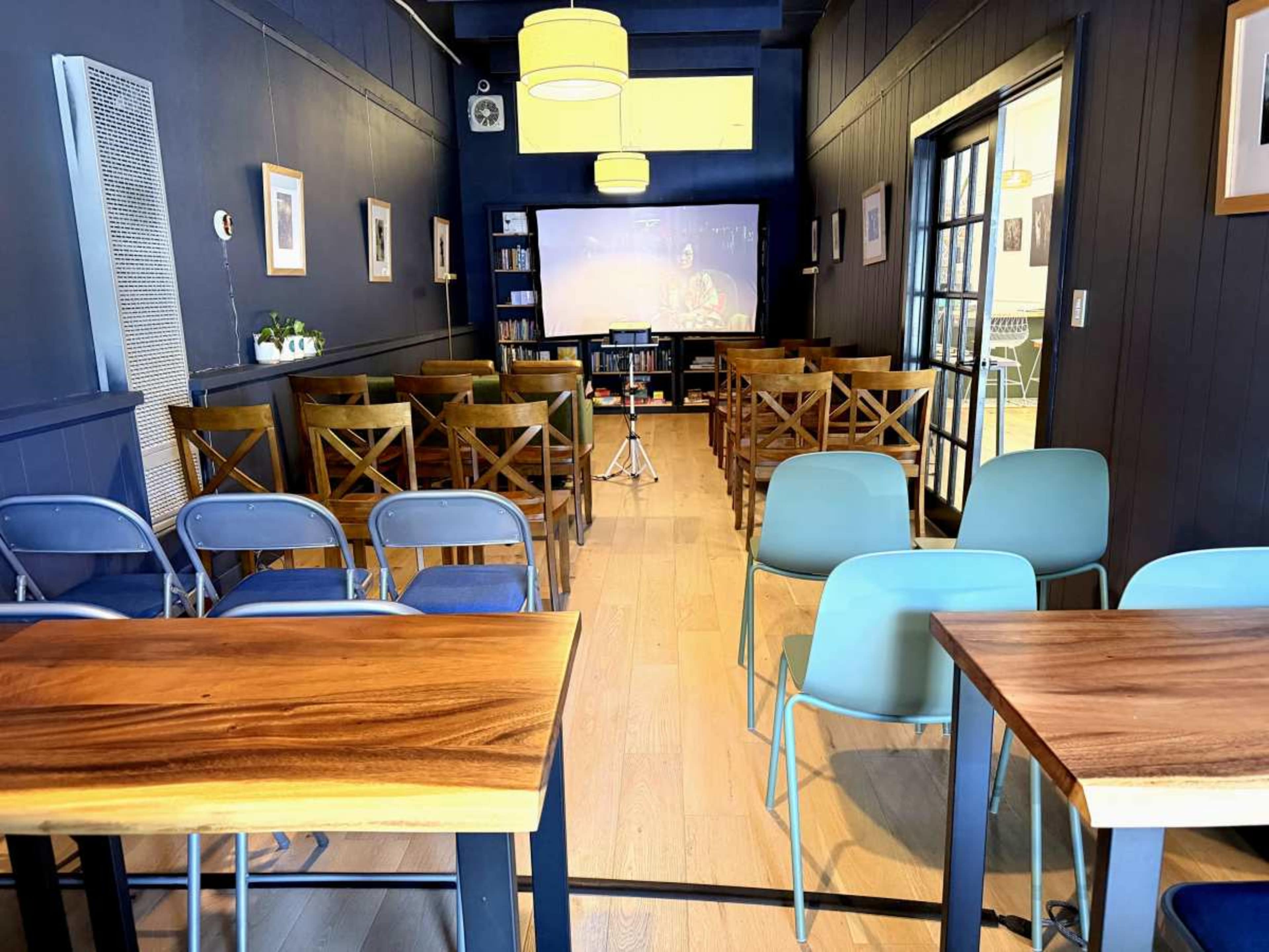 The image shows a neatly arranged indoor space with chairs and tables, featuring a screen in front and shelves filled with books on one side.