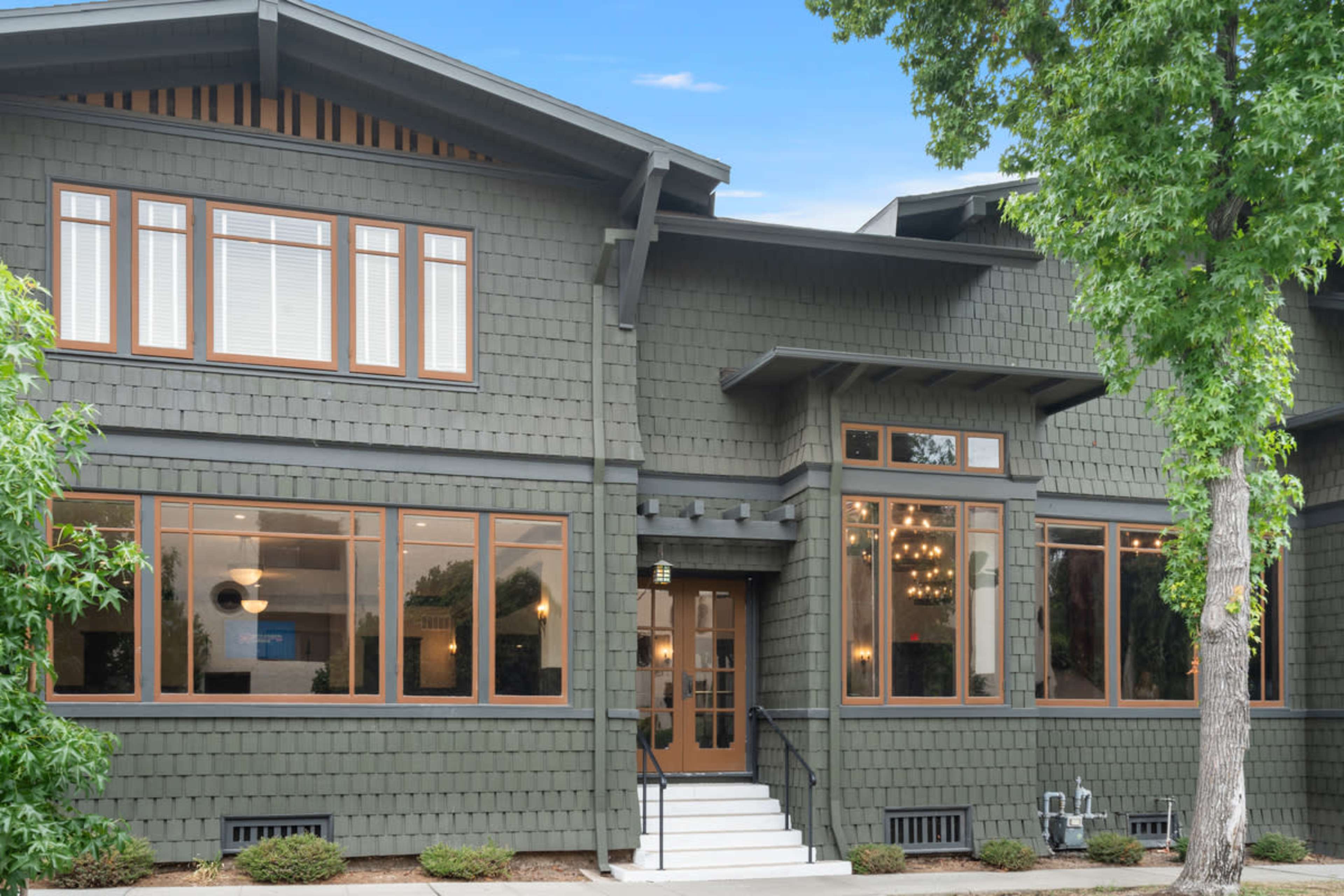 The image shows a large, multi-story house with dark green shingle siding, multiple windows, and a front entrance featuring stairs and a light fixture.