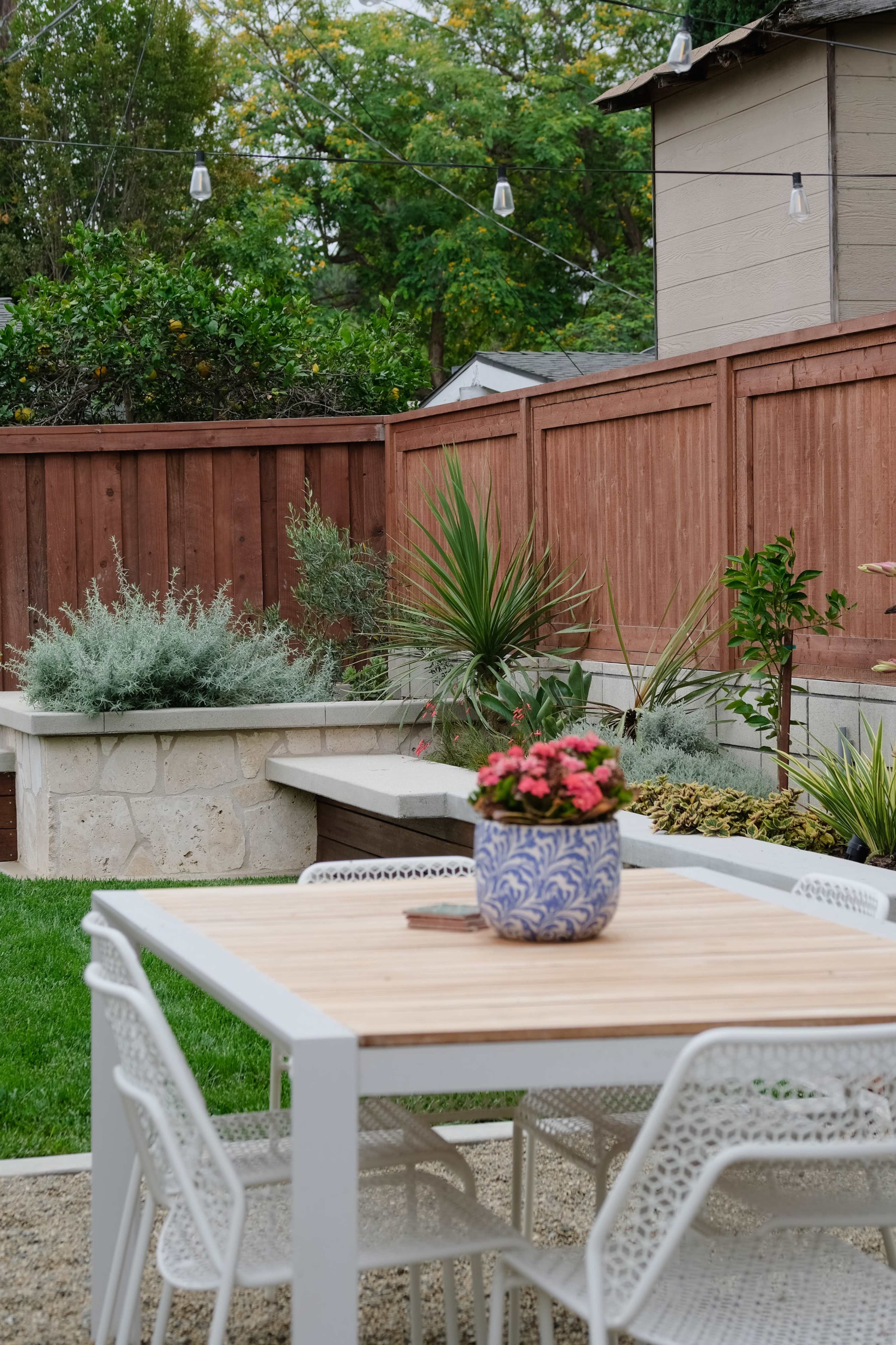 A modern outdoor dining area features a wooden table surrounded by white chairs, with a potted plant and landscaped greenery in the background.