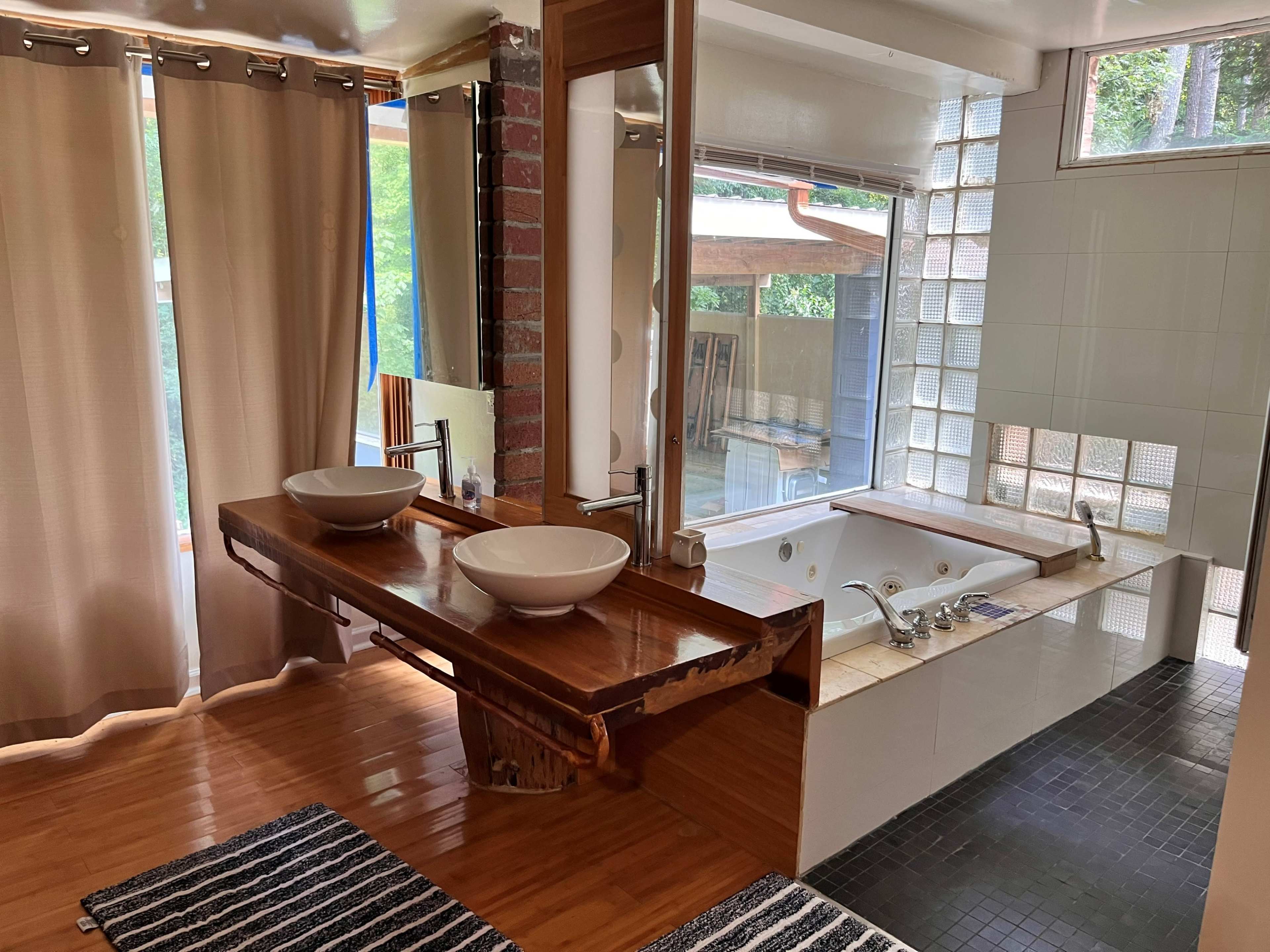 A modern bathroom featuring a wooden vanity with two sinks, large mirrors, and a spacious area with a jacuzzi tub and a glass block wall.