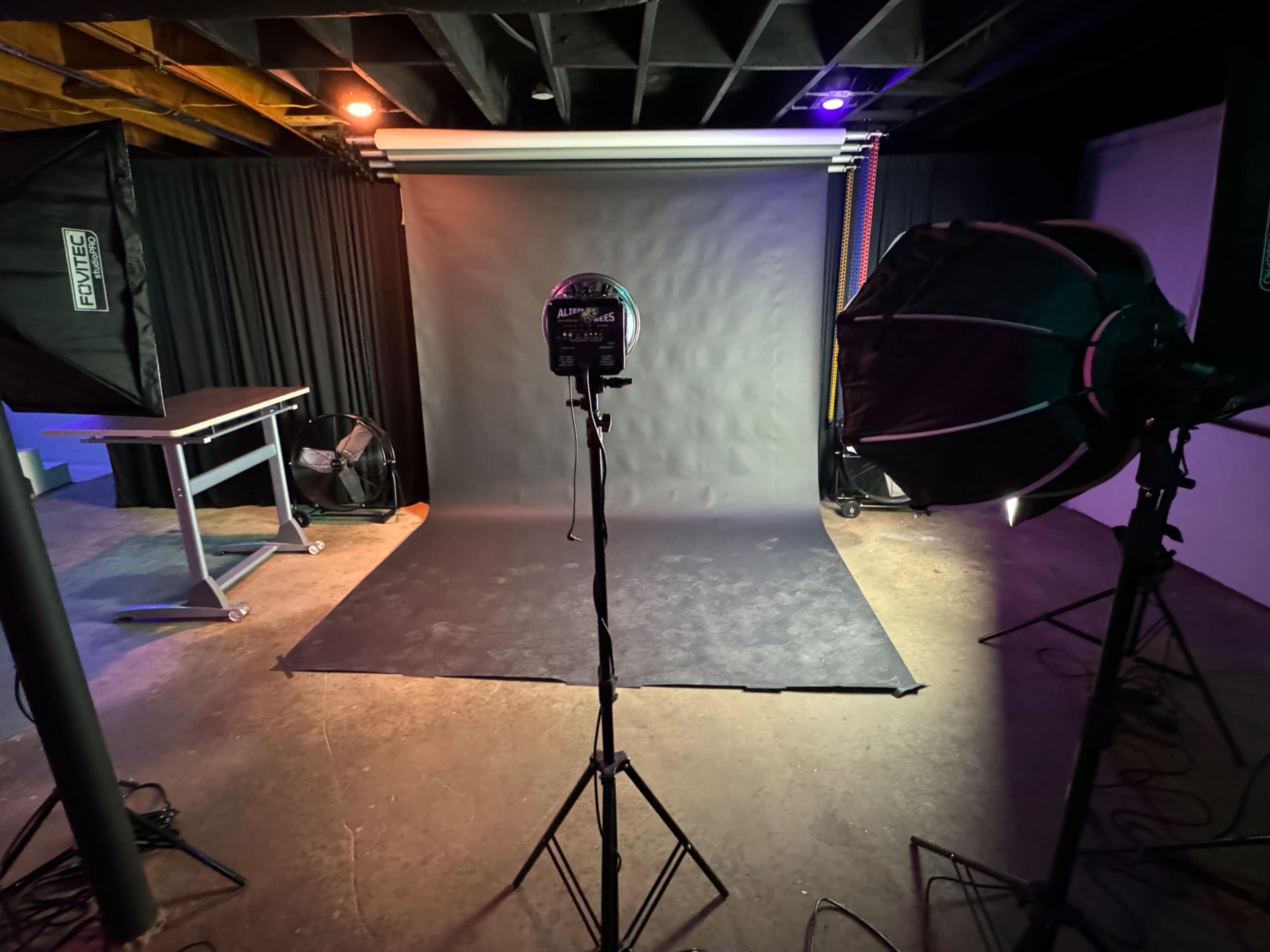 A photography studio setup featuring a black backdrop, two lighting softboxes, and a tripod for a camera.