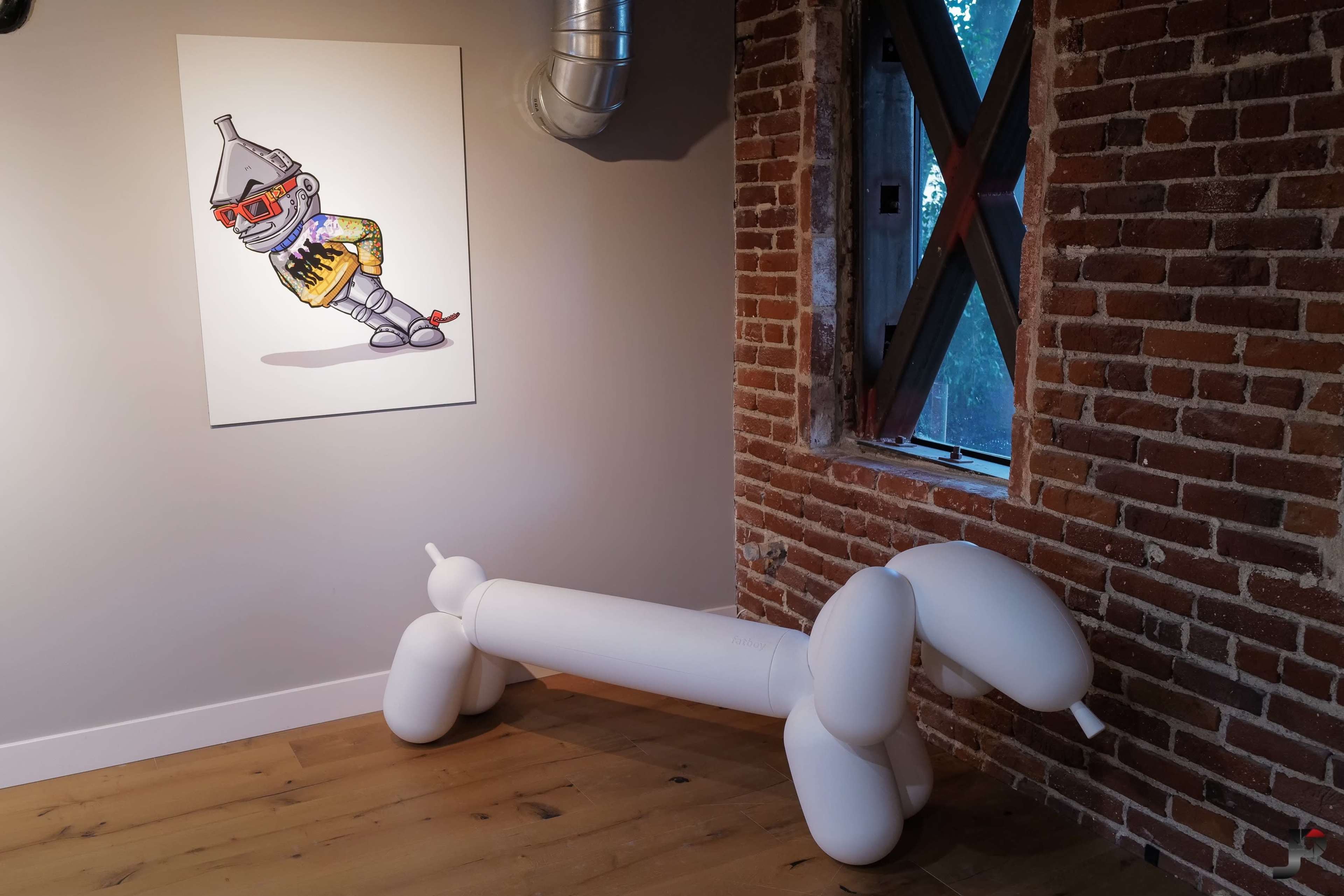 A balloon sculpture resembling a dog sits on the floor next to a framed painting of a cartoon character in an industrial-style room with exposed brick walls and a window.