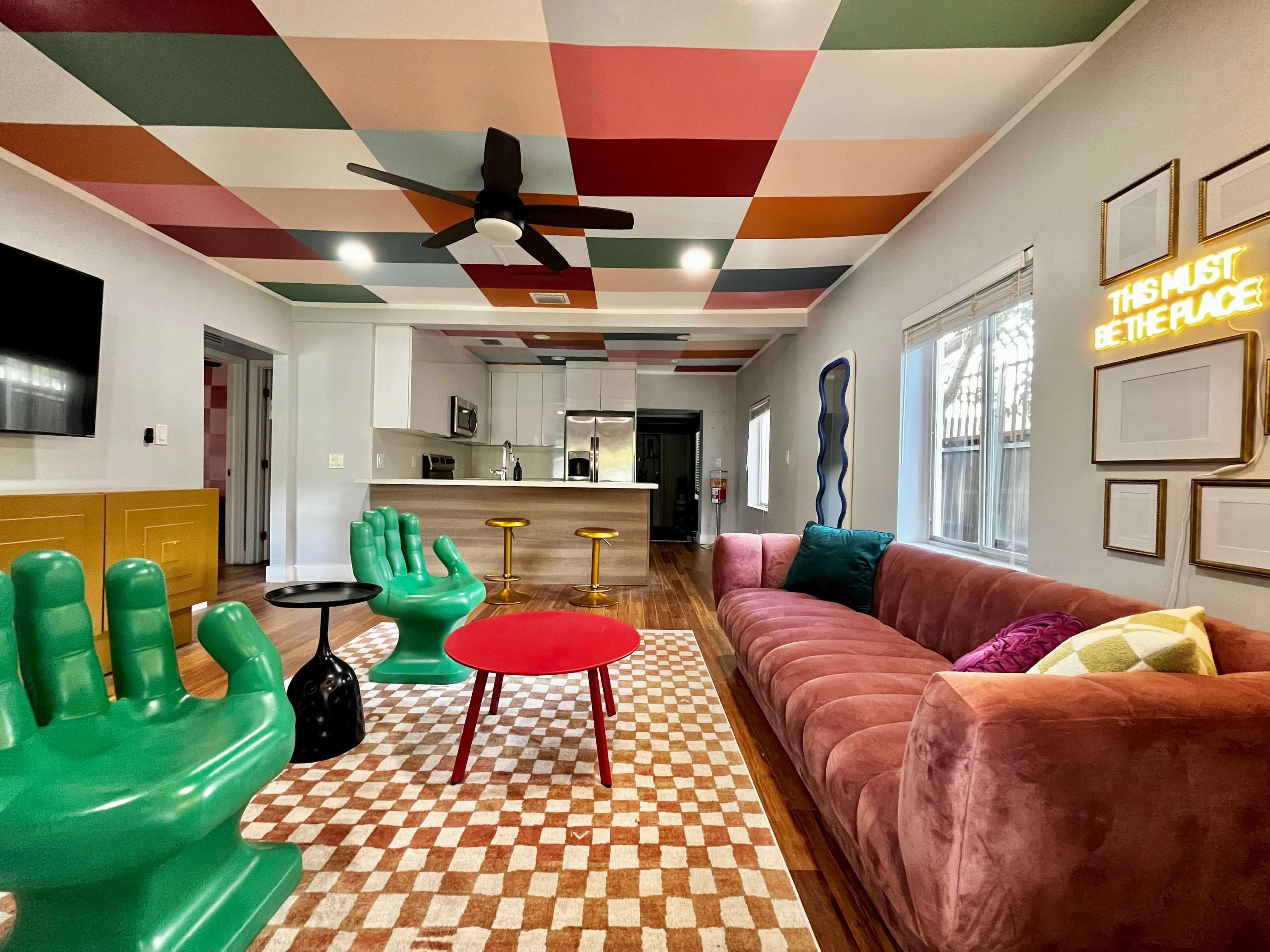 The room features a colorful ceiling, a pink sofa, green hand-shaped chairs, and a red coffee table on a patterned rug.