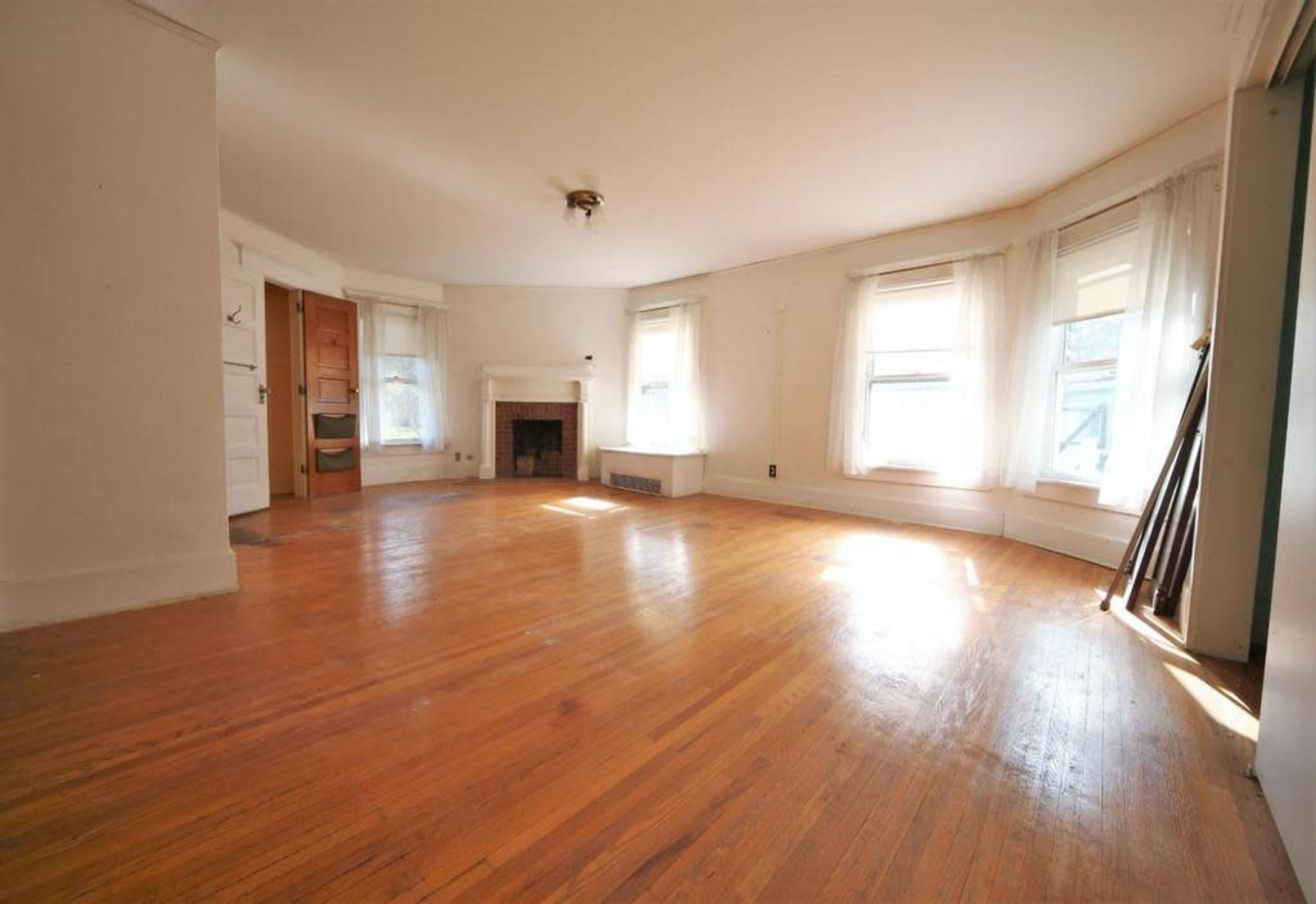 A bright, empty living room with hardwood floors, large windows, and a fireplace.