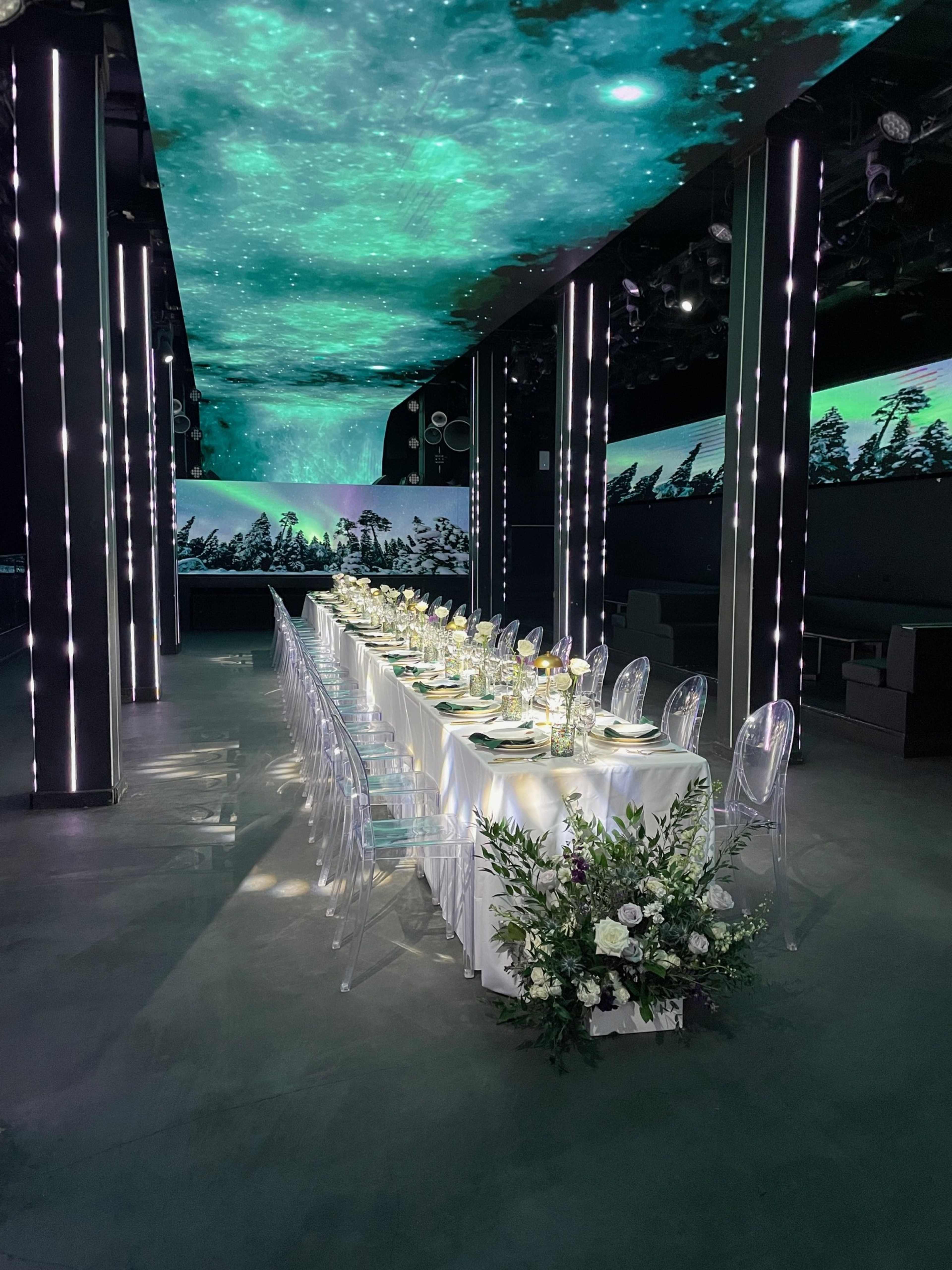 A long dining table is set with plates and glasses under a ceiling projection of a starry night sky, surrounded by greenery and modern decor.