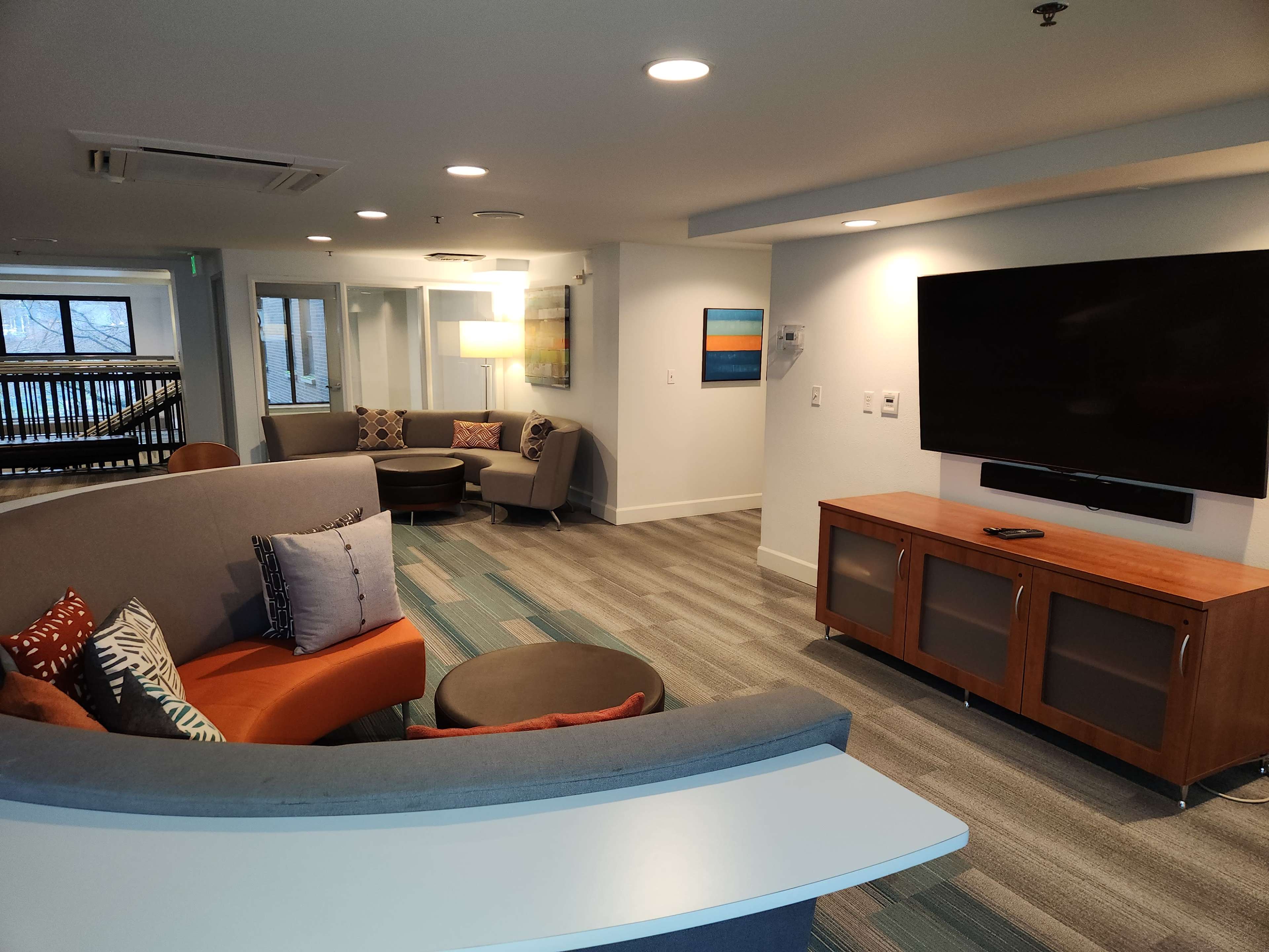 A modern lounge area featuring a curved gray couch, a wooden television cabinet, and colorful cushions, with a flat-screen TV mounted on the wall.