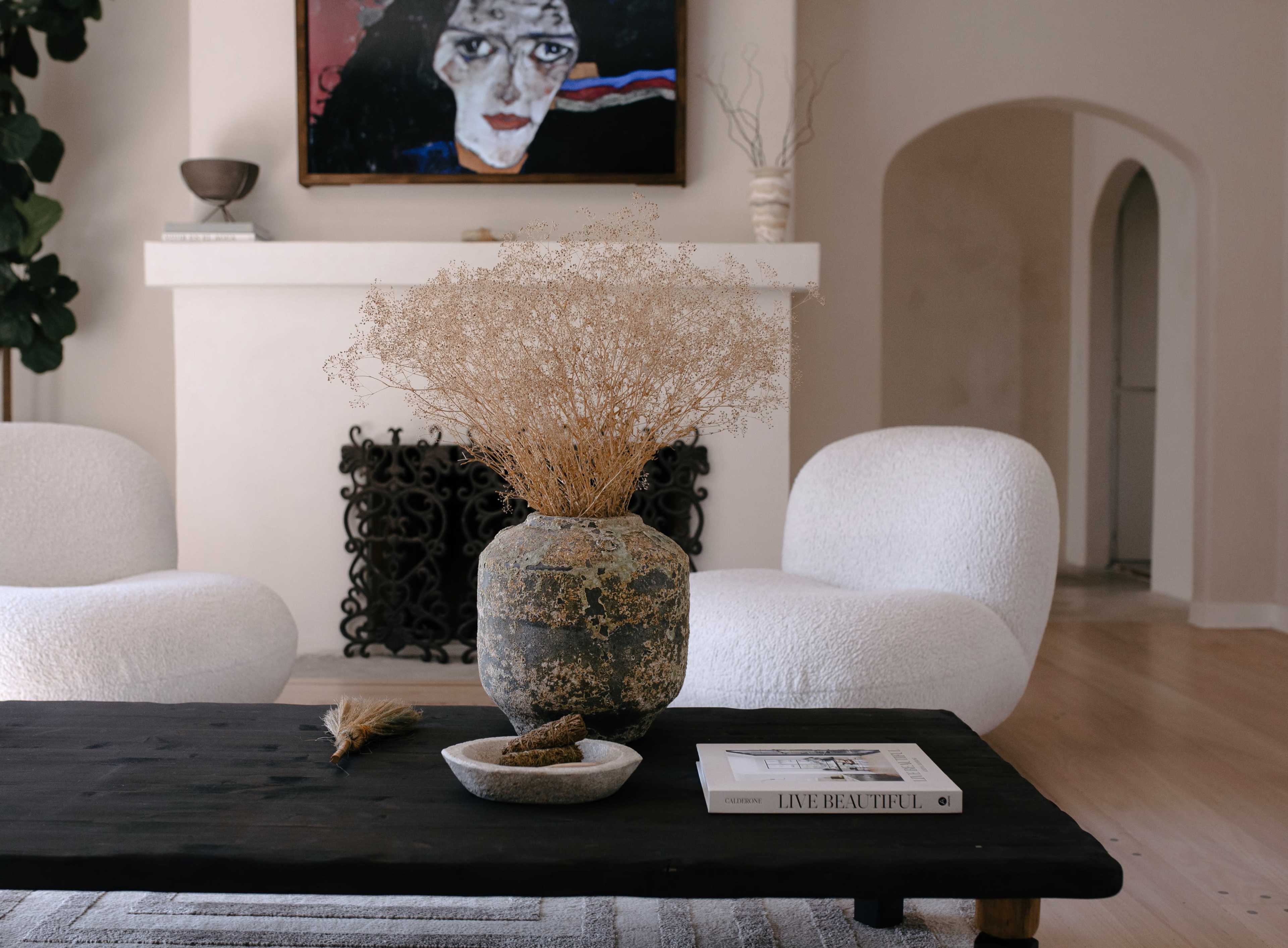 A rustic coffee table holds a textured vase filled with dried flowers, paired with a small dish and a book, surrounded by white modern chairs in a minimalistic living space.