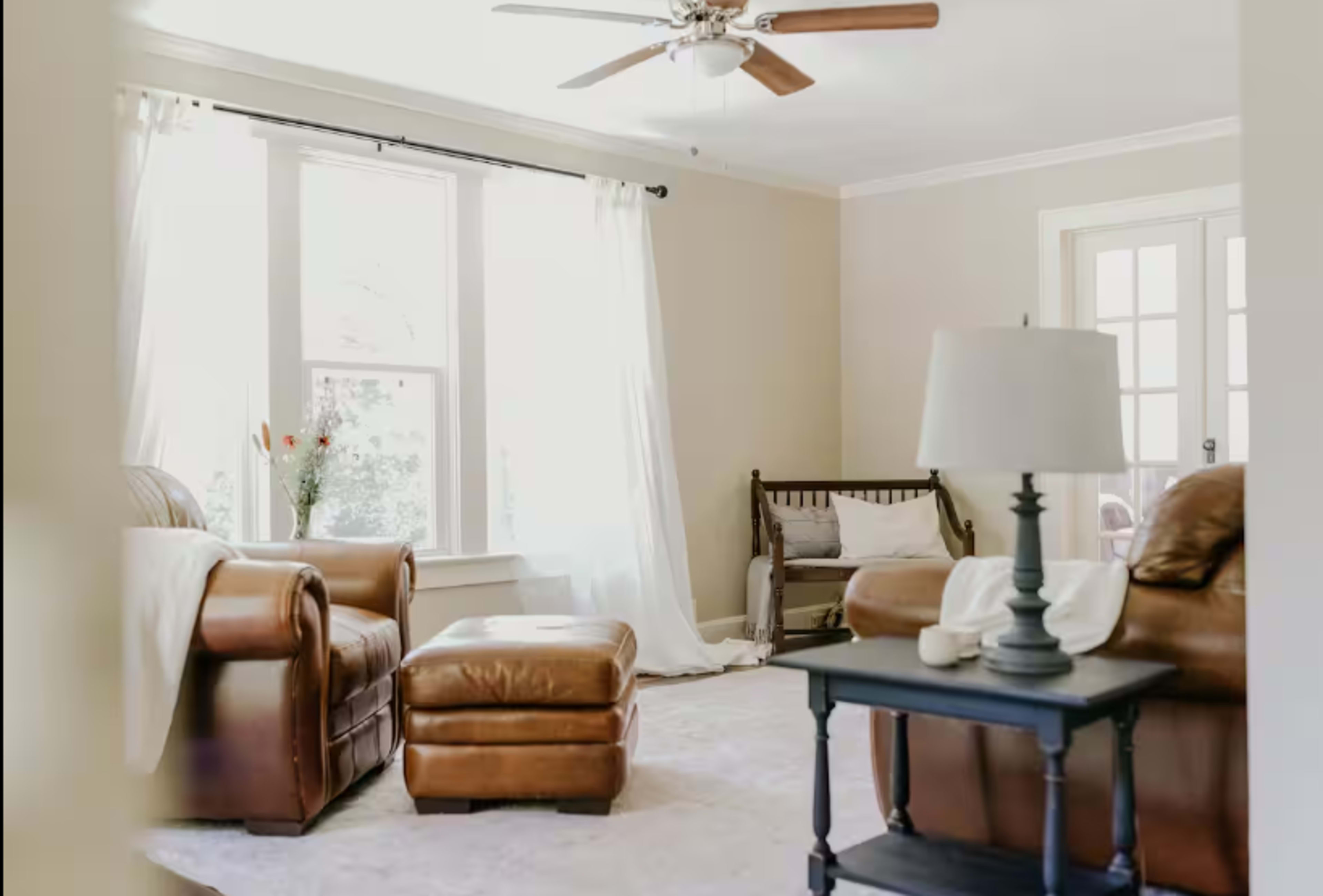A living room features two brown leather sofas, a matching ottoman, a table lamp, and large windows with sheer curtains allowing natural light to enter.