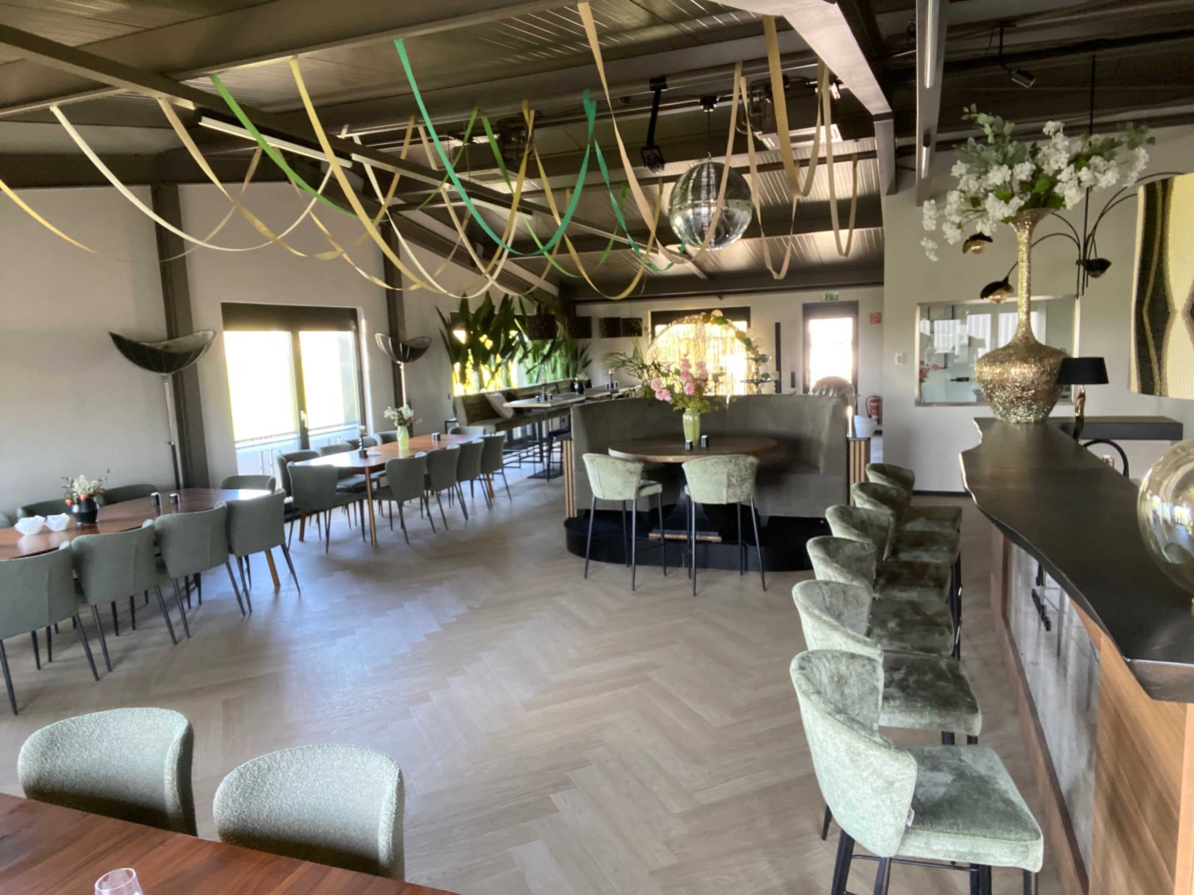 The image shows a modern restaurant interior featuring neatly arranged tables, green chairs, and decorative elements such as hanging ribbons and plants.