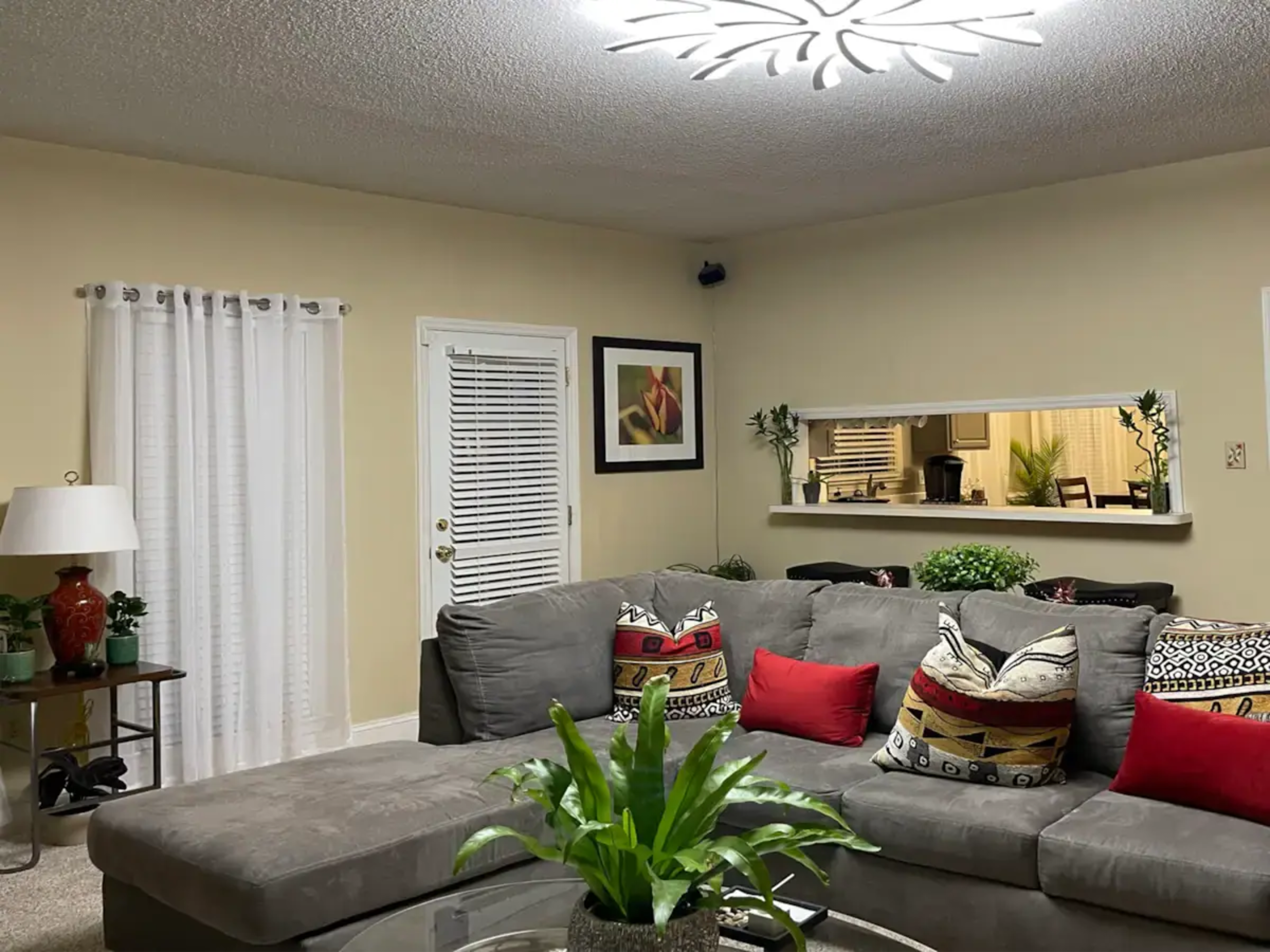 A cozy living room with a gray sectional sofa adorned with colorful pillows, a round glass coffee table, and a wall-mounted light fixture above.