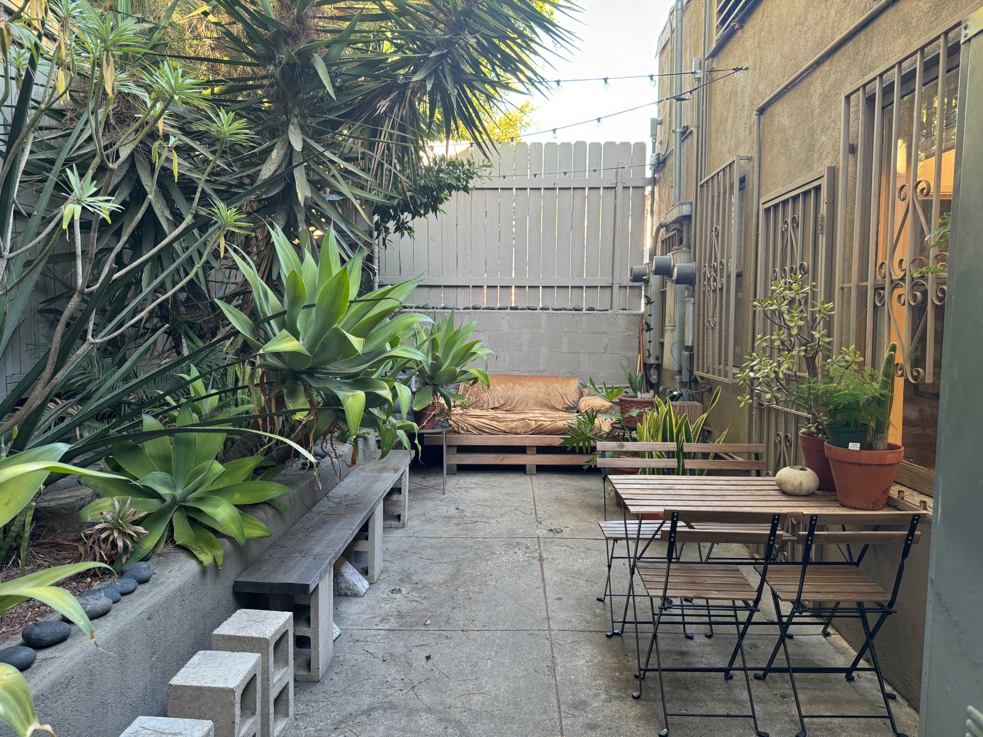 A compact outdoor patio with a couch, potted plants, and seating made from small tables and blocks, bordered by walls and a fence.