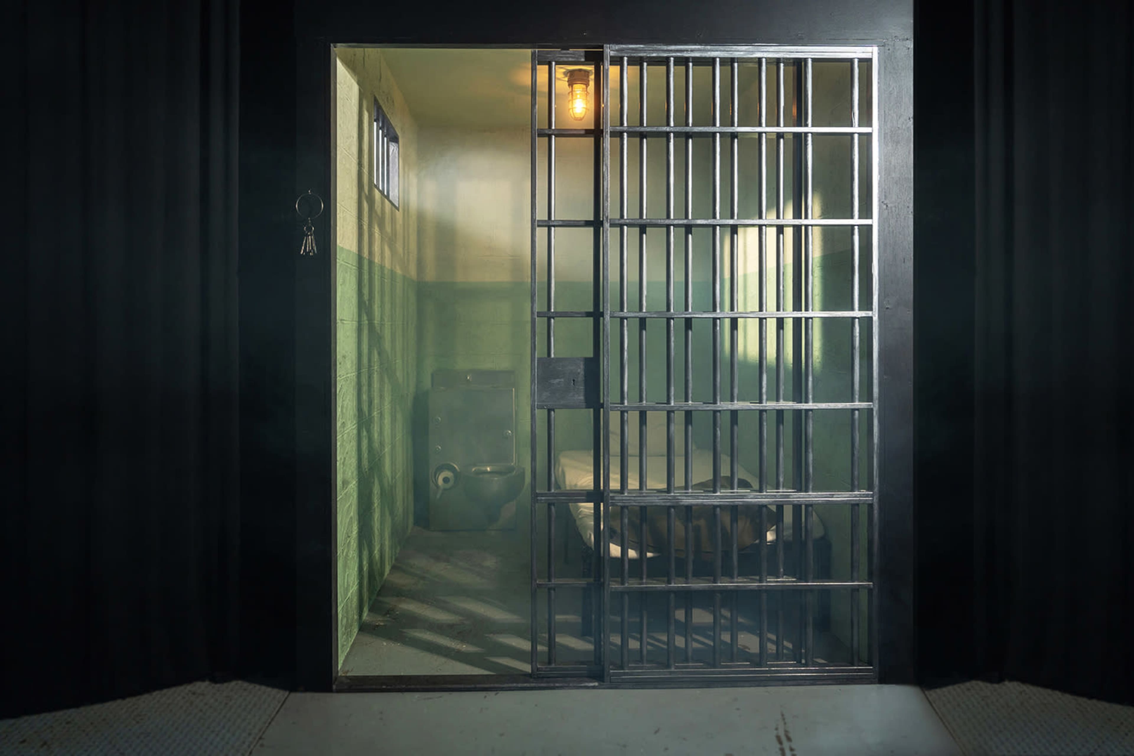 The image shows the interior of a prison cell with a barred door, a bed, a toilet, and a single light bulb hanging from the ceiling.