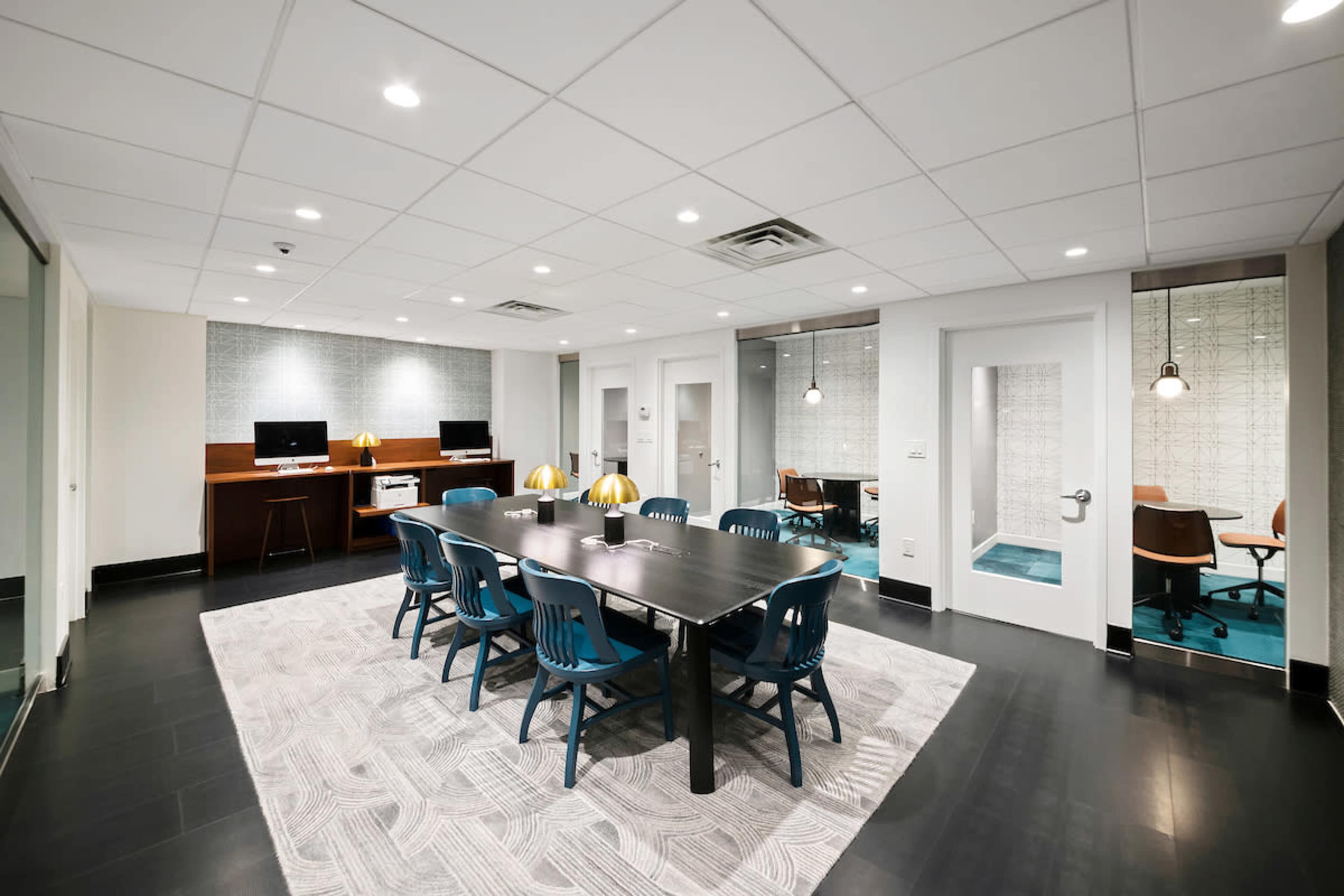 The image shows a modern conference room featuring a large rectangular table surrounded by blue chairs, with two workstations and glass-walled meeting rooms in the background.